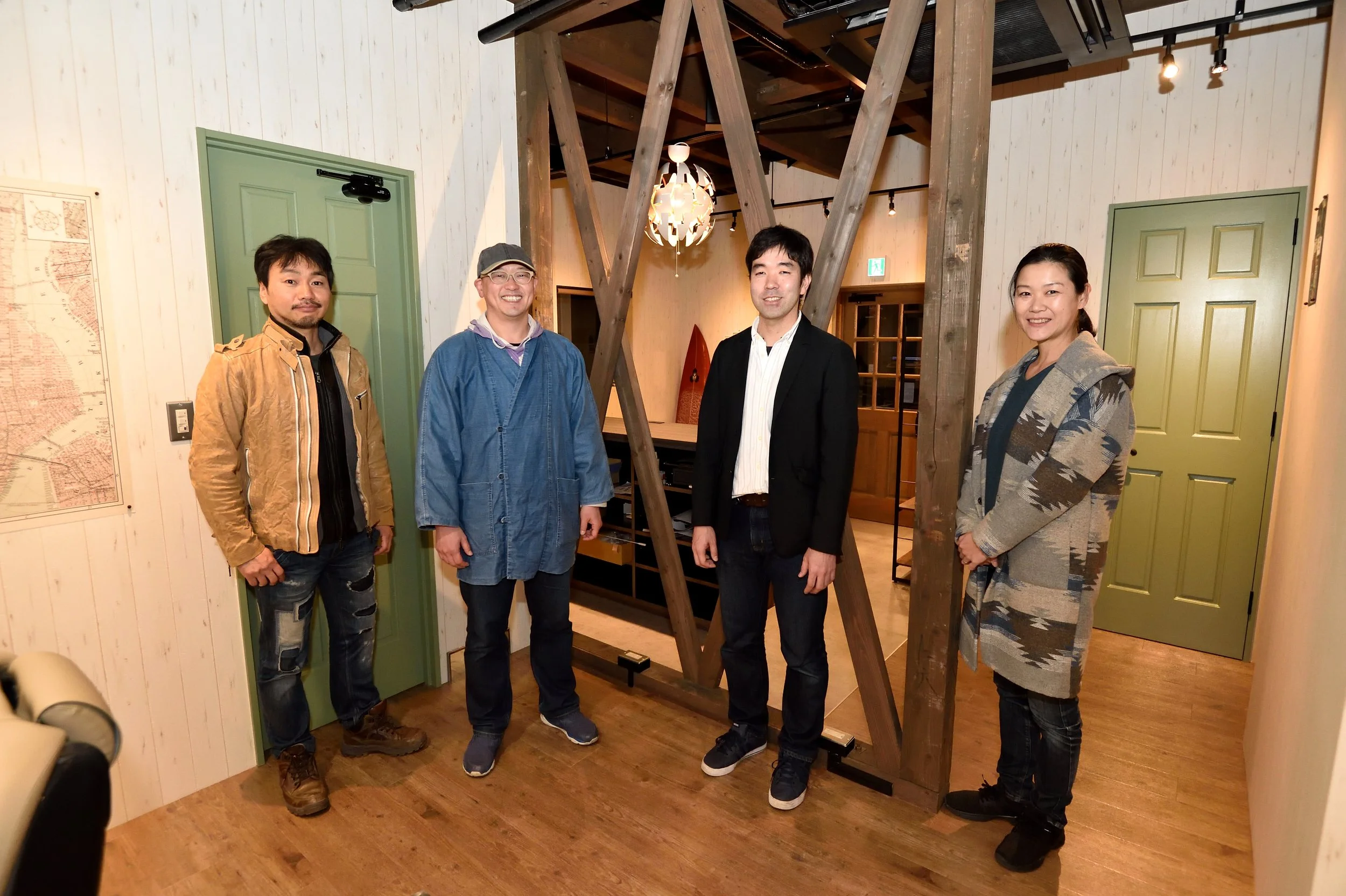 木の梁を背景にした室内で、4人の人々が笑顔で並んでいる。左から男性、男性、男性、女性。壁には地図が掛かっている。
