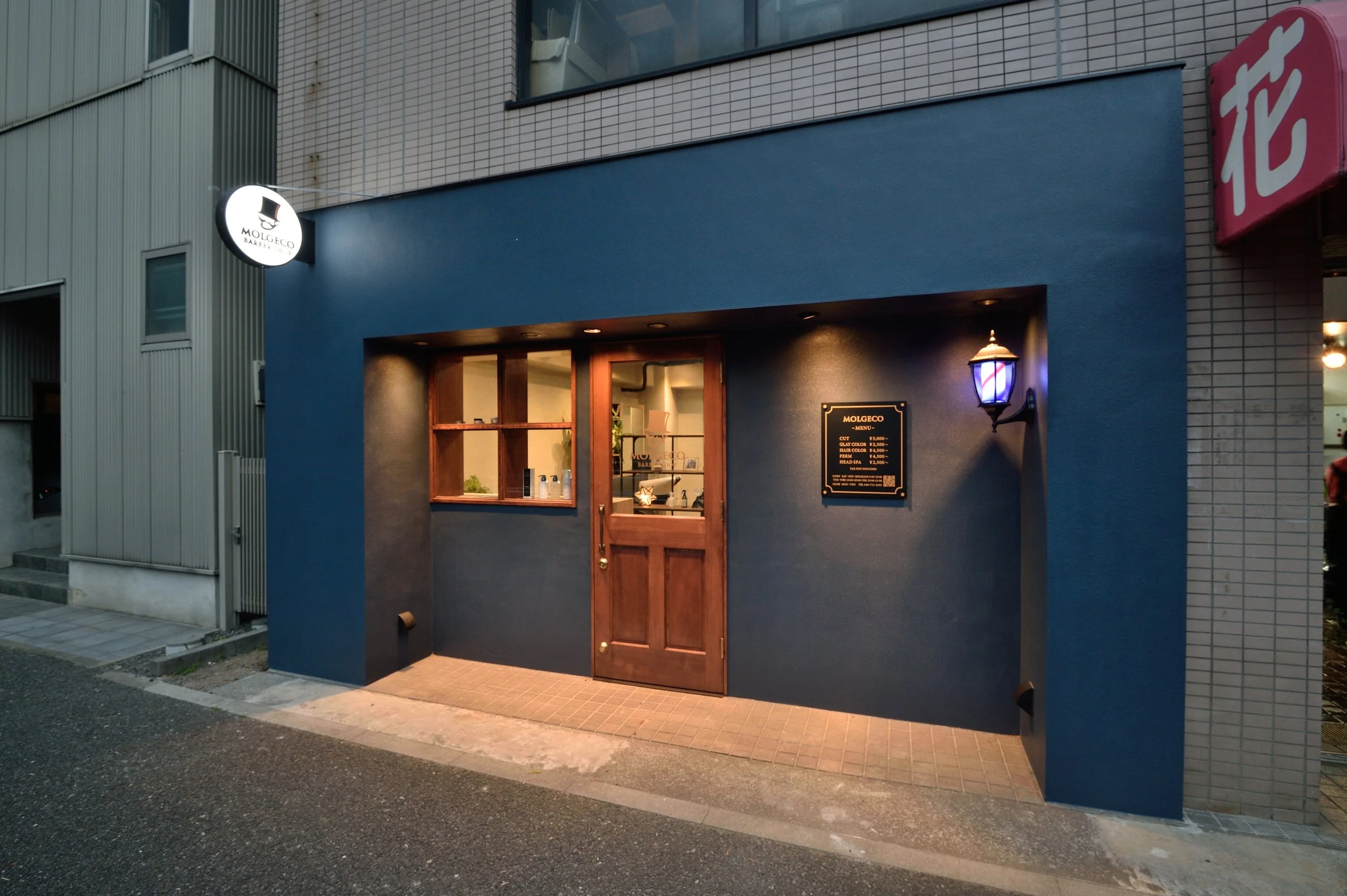 青い外壁の建物の入り口に木の扉と窓、扉の横に看板とライトが設置されている。