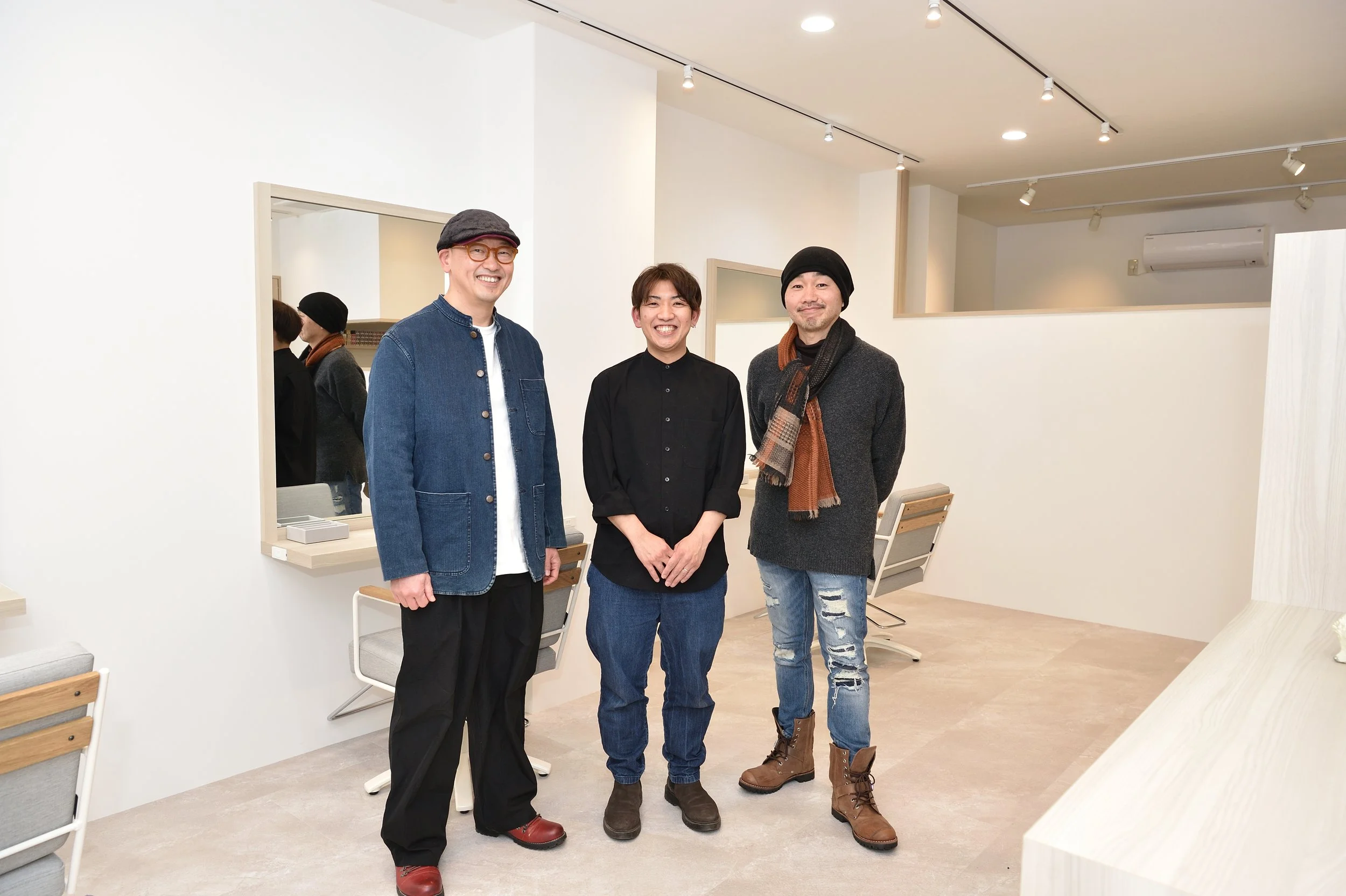 白い壁の部屋で、中央に三人の男女がお辞儀をして笑顔で立っている。背後には鏡と椅子が見え、天井にライトが設置されている。