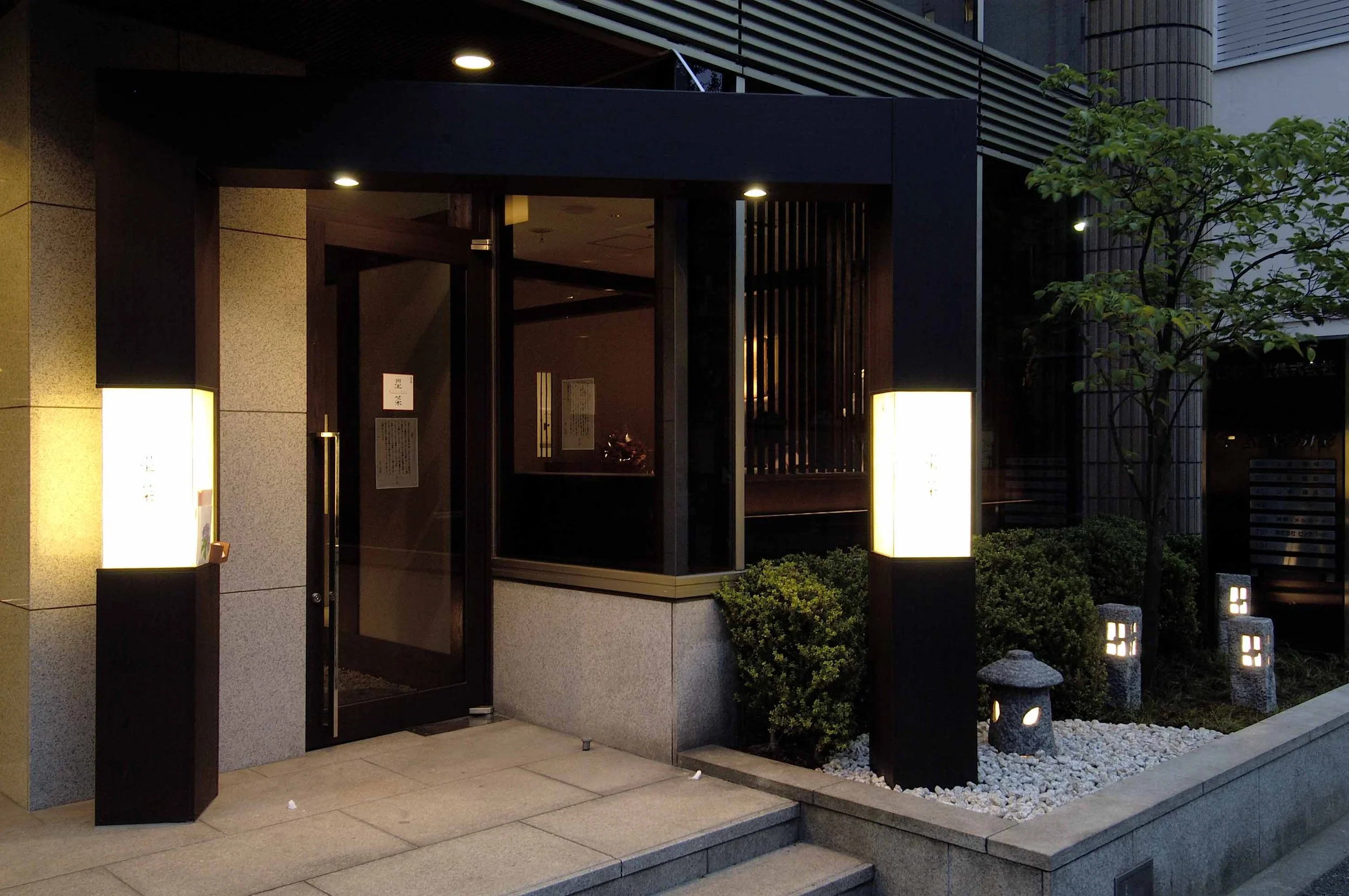 夜のエントランスの入口、照明付きの立て看板と植物があるモダンな建物の外観