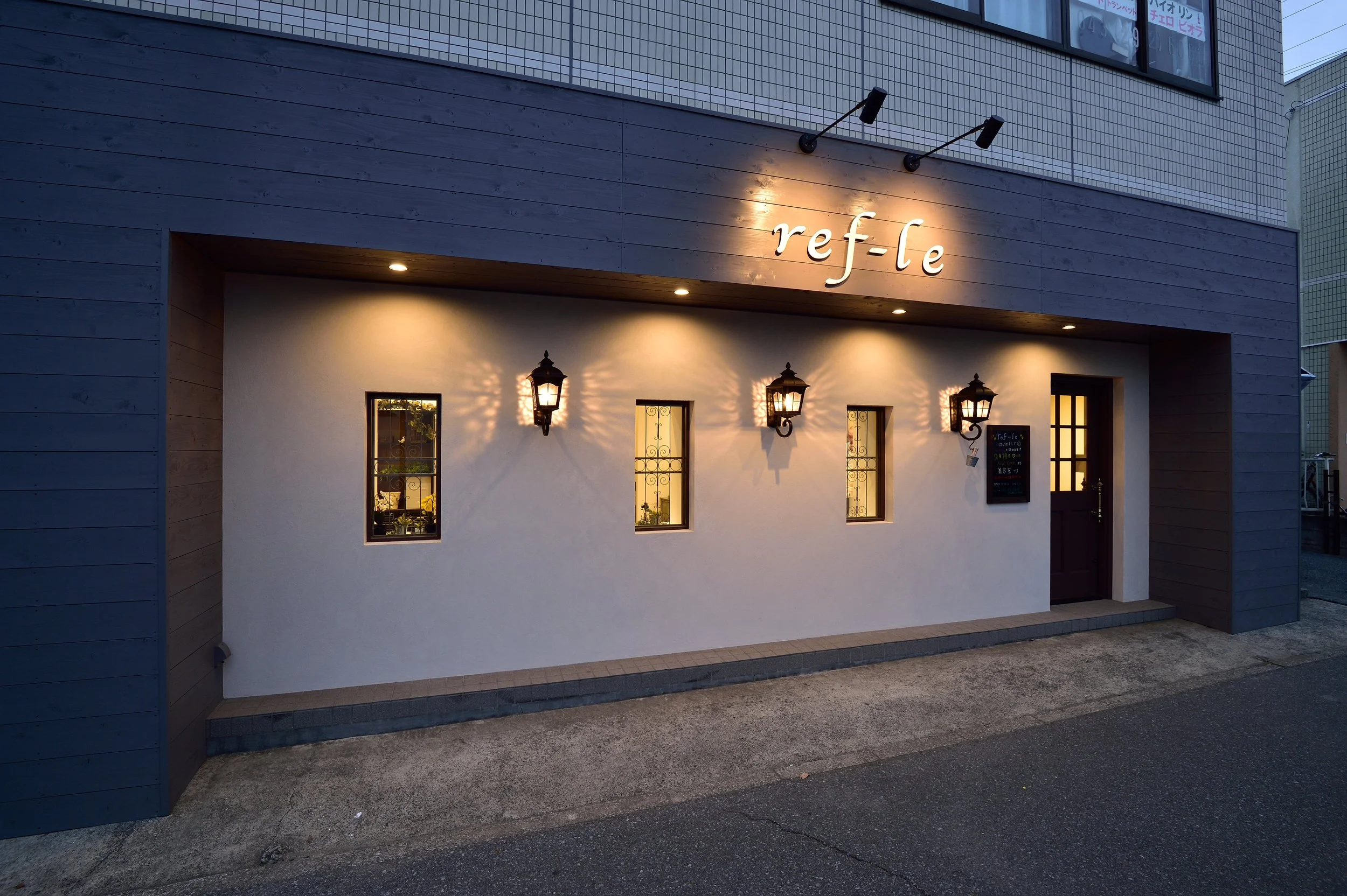 建物の外観、白い壁と木製の看板に「ref-le」のサインがあり、暖かい照明のブラケットライトが壁に設置されている。夜のような暗い時間帯の写真。