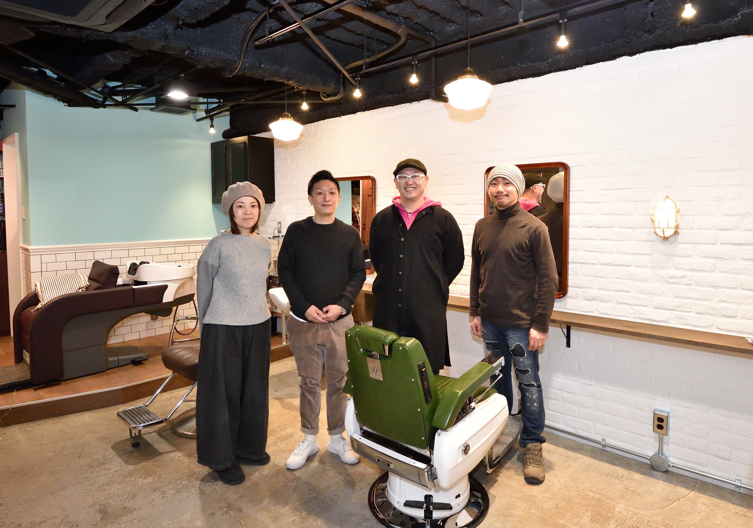 室内で4人の人々がカメラに向かって笑顔で立っている。背景には白いレンガの壁と鏡があり、天井には黒い配管と照明が見える。