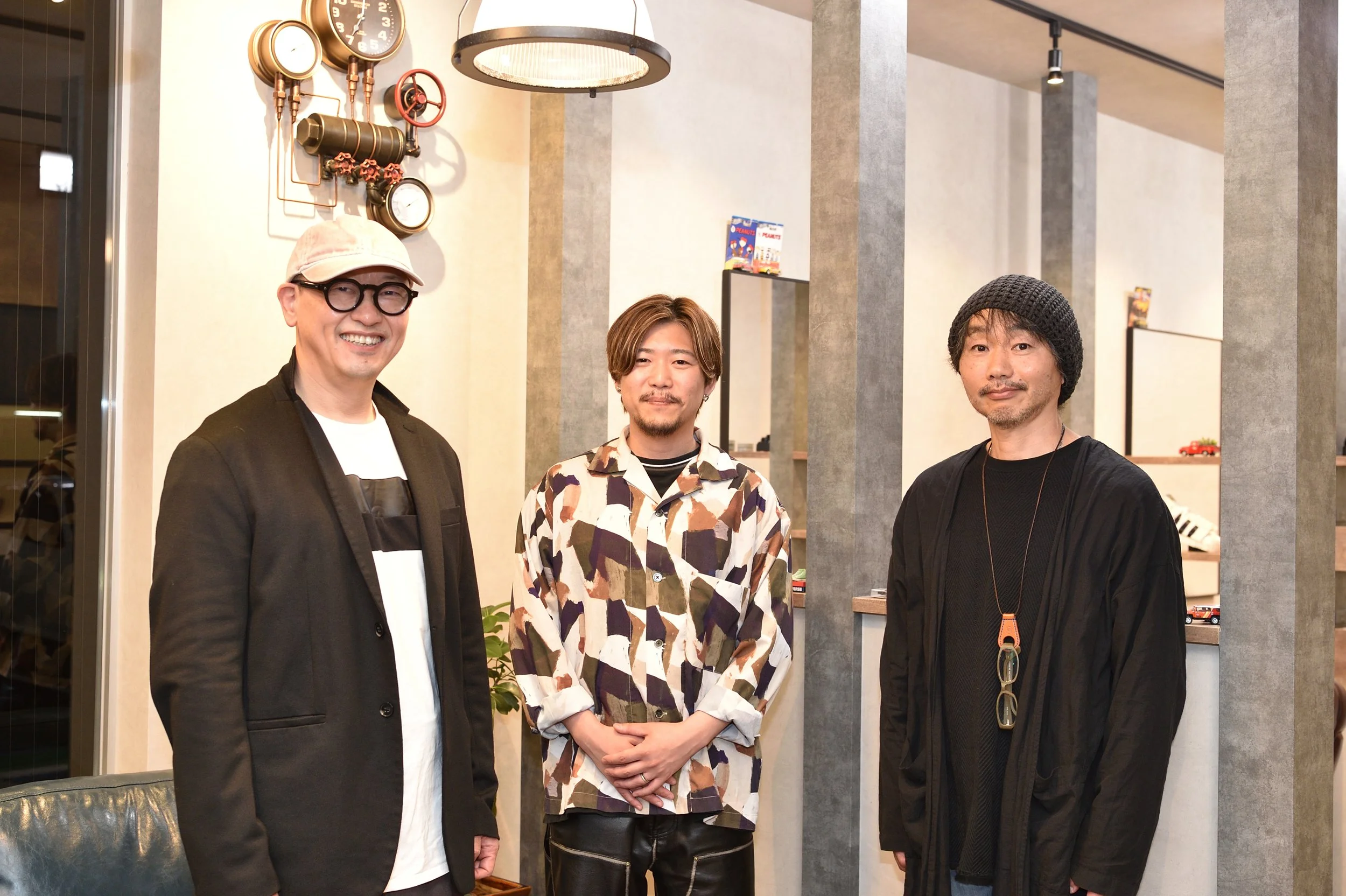 店舗開業ｲﾝｽﾄﾗｸﾀｰｂｈ飯島由敬と開業者の店舗OPEN時の記念撮影写真。カメラの前で笑顔の3人の男性が室内に立っている。背景には壁掛けの時計や装飾が見える。男性はカジュアルかつスタイリッシュな服装をしている。