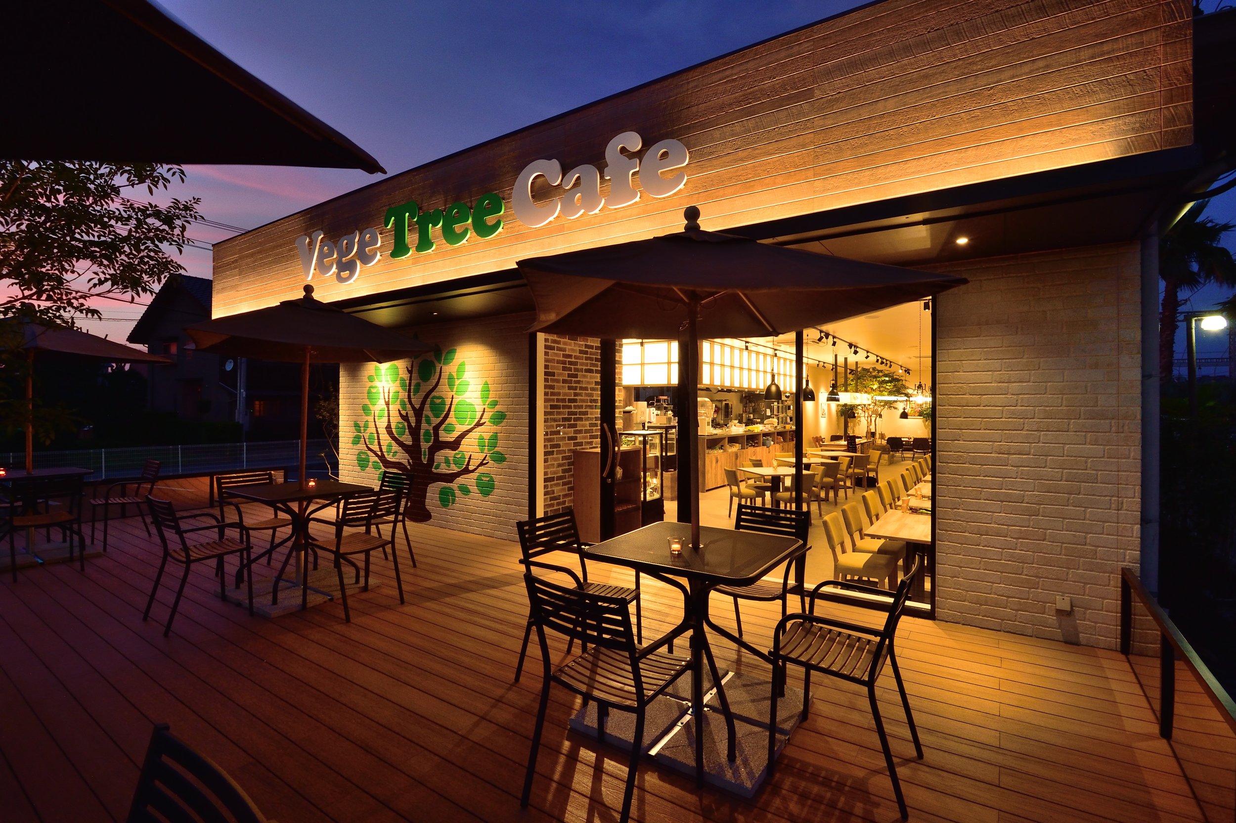 木材の外観のカフェの看板が夜に照明に照らされている画像。屋外のテラス席にはテーブルと椅子が並び、天気の良い夕暮れの空が背景に見える。