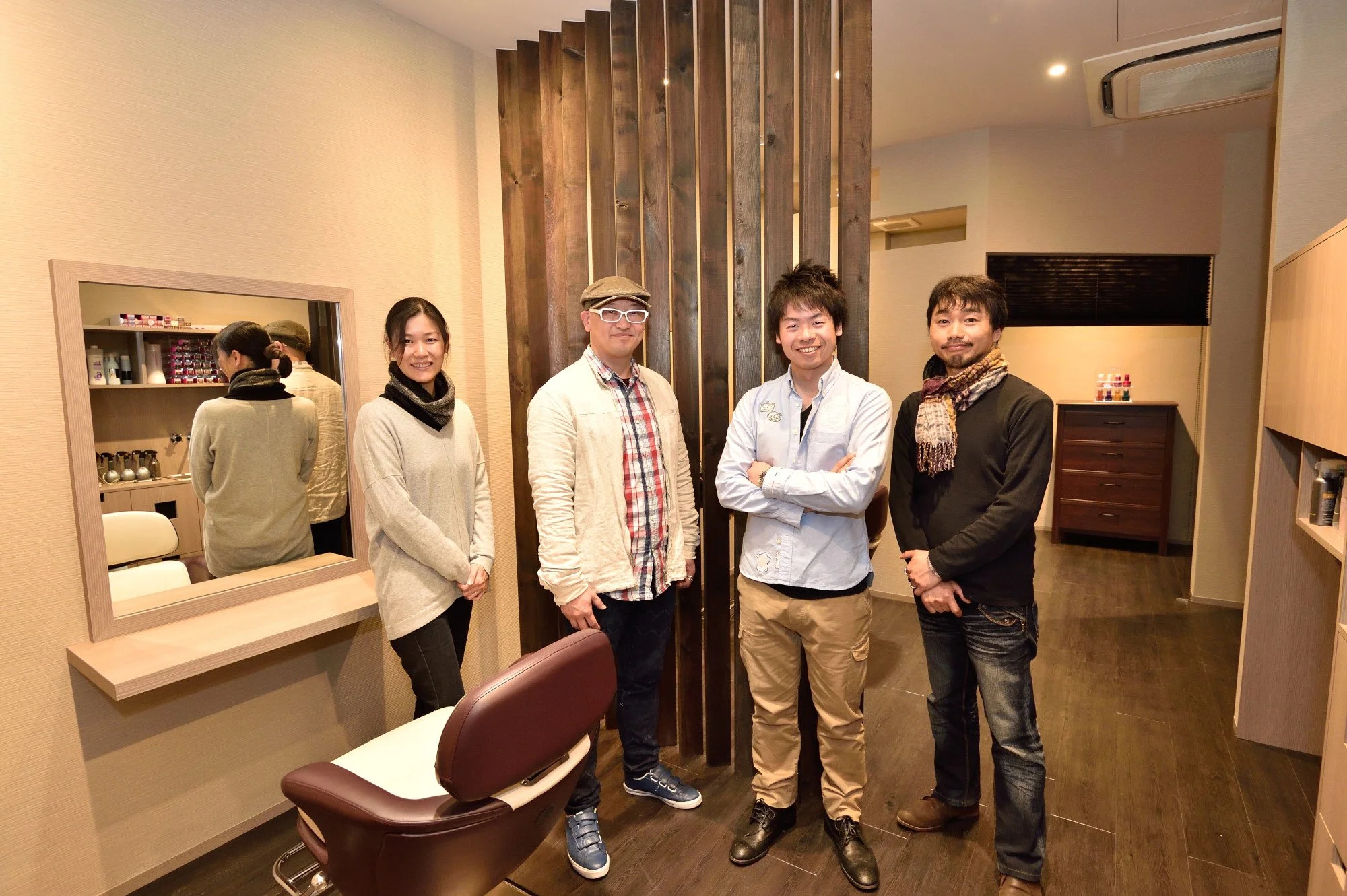 四人の男女が室内で笑顔で立っている。背景には木製の仕切りと鏡や棚がある。