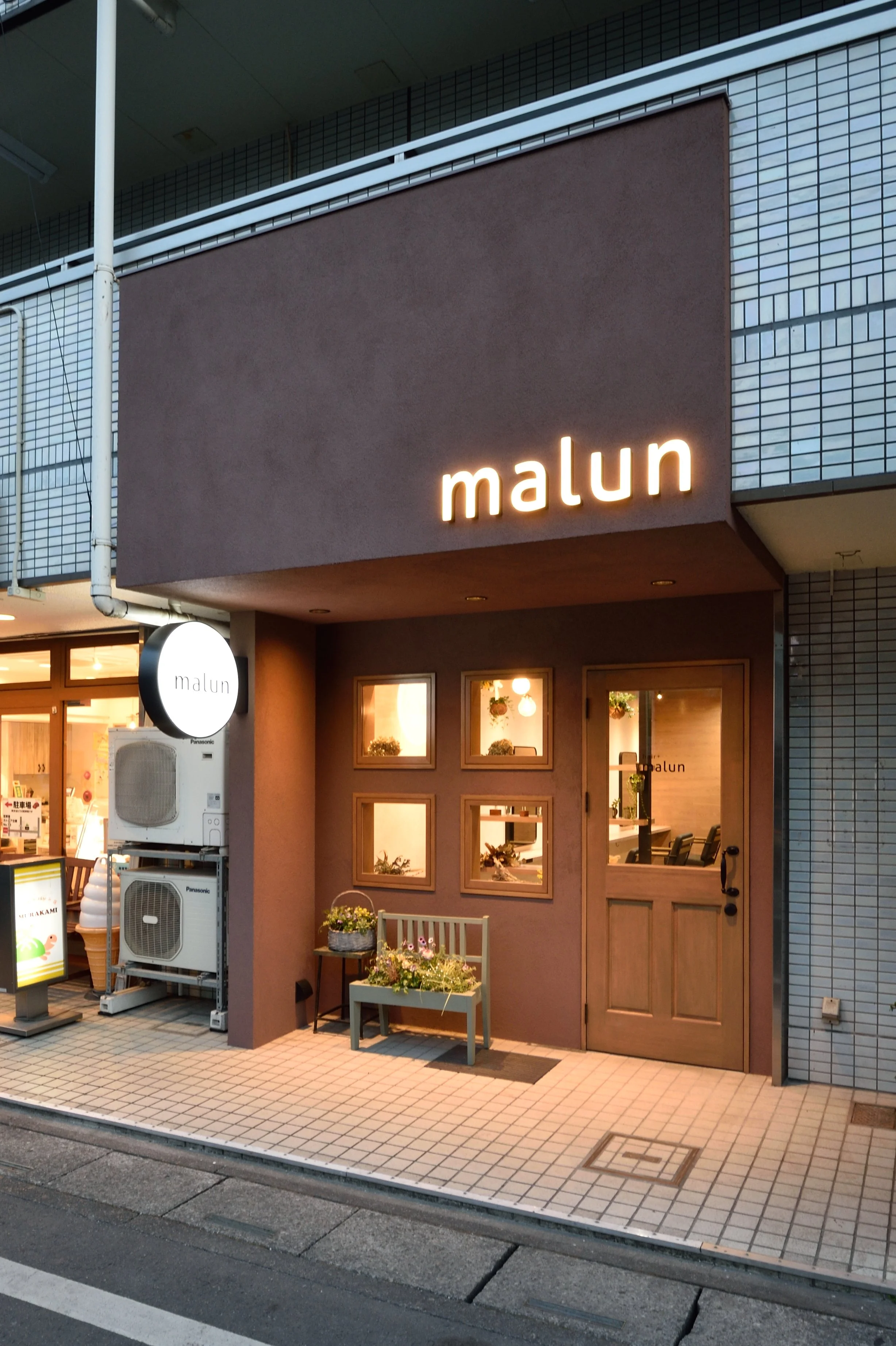 シンプルなデザインのカフェやレストランの外観。看板には「malun」と書かれている。入口には窓があり、中に花の飾りが見える。敷地には小さなベンチと花の鉢が置かれている。