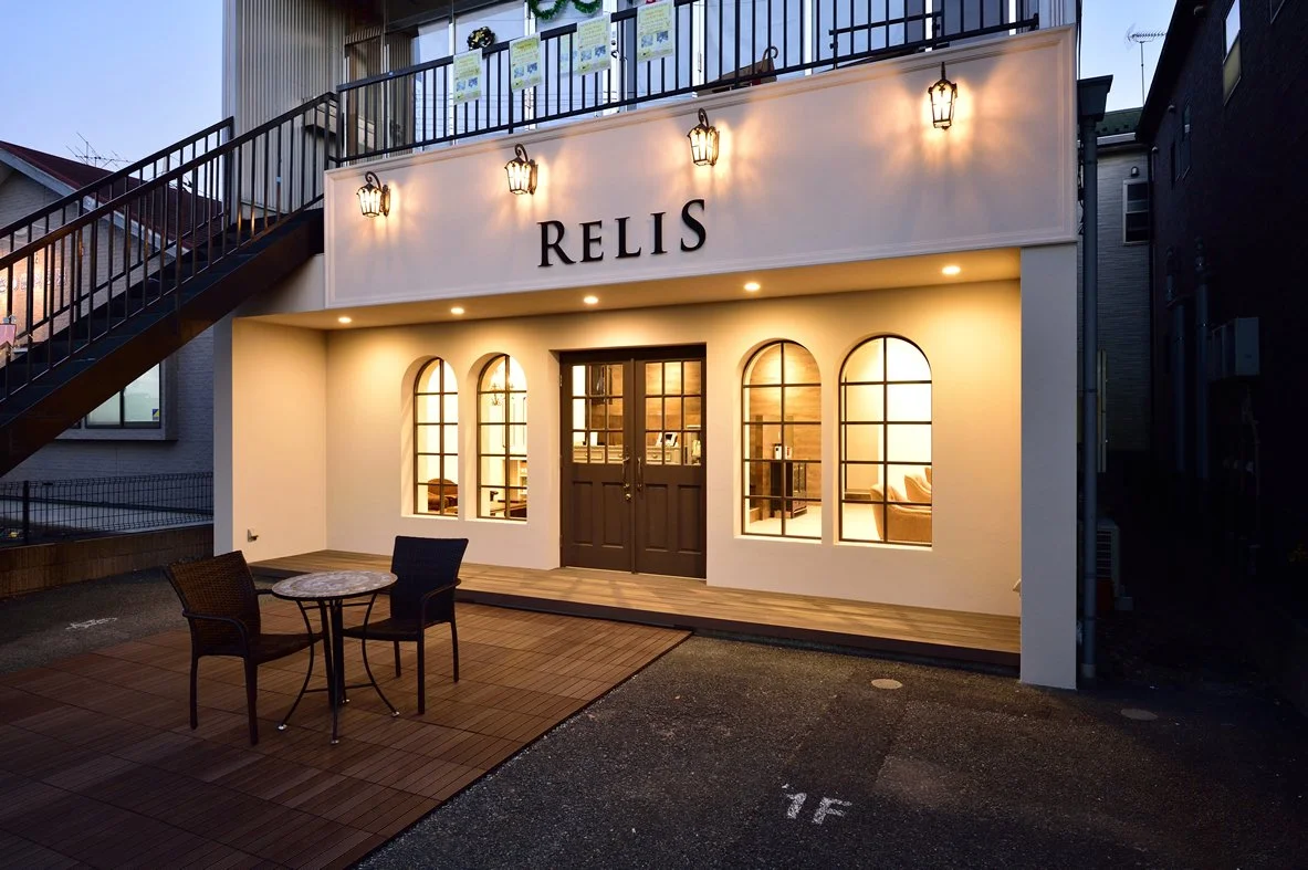 夜間のカフェの外観。白い壁に黒い文字で"RELIS"と書かれており、窓から温かい光が漏れている。外にはテーブルと椅子があり、階段が左側にある。