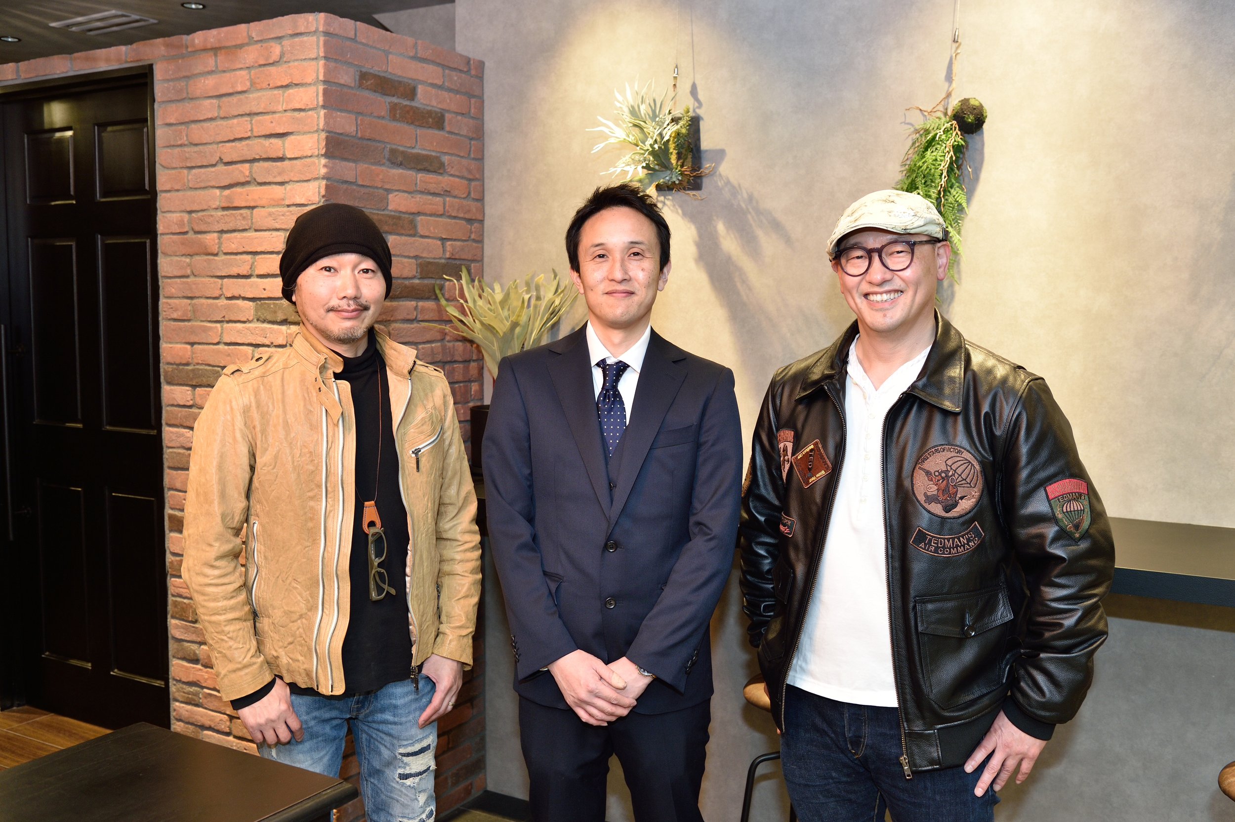 店舗開業ｲﾝｽﾄﾗｸﾀｰｂｈ飯島由敬と開業者の店舗OPEN時の記念撮影写真。スーツ姿の男性とカジュアルな服装の男性2人が笑顔で並んでいる。背景にレンガの壁とグリーンの装飾がある。