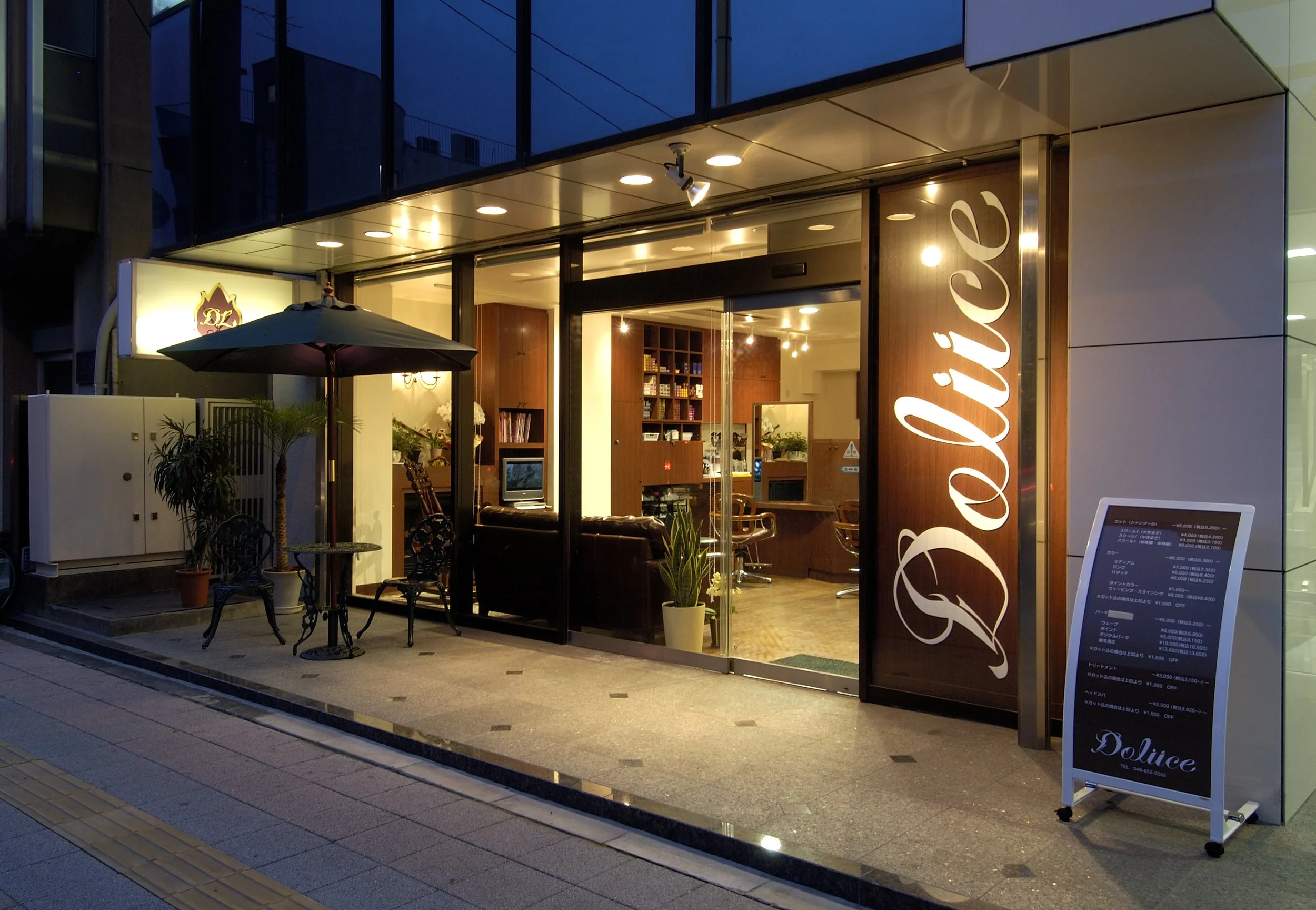 夜の外観の美容室の入り口。ガラス窓越しに内装が見え、看板には「Dolice」と書かれている。外には黒いパラソルとチェア、メニューの立て札が置かれている。