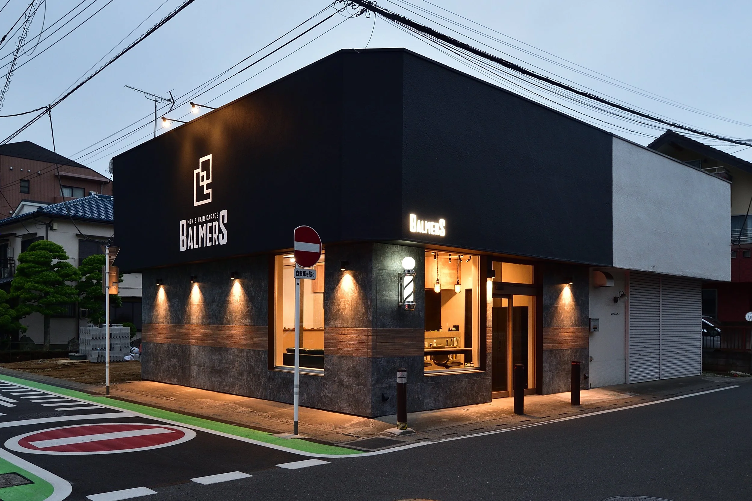 暗くなる前の夕暮れ時、建物の外観に「BALMERS MEN'S HAIR GARAGE」の看板がある美容院の店内の灯りがついている。
