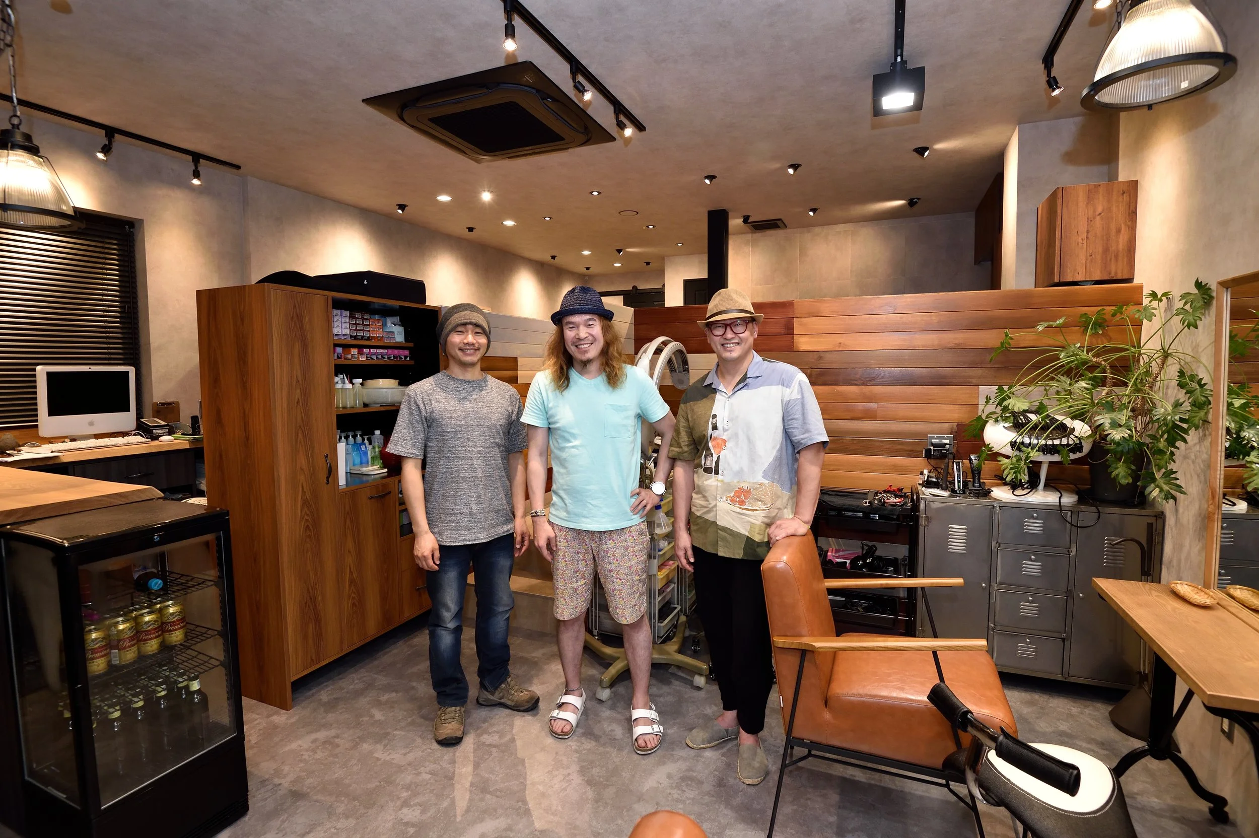 室内のカフェまたはレストランで、中央に3人の男性が笑顔で立っている。背景には木製の壁と植物、電子機器や収納棚が見える。