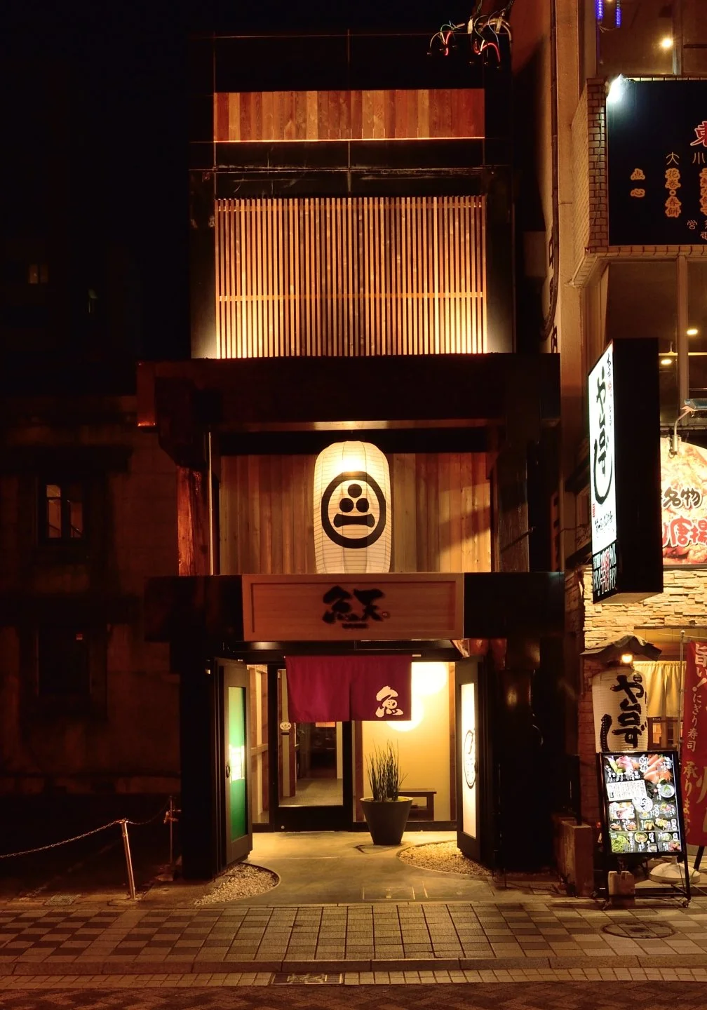 夜の日本のレストランの入口に提灯と看板が飾られている。木造の壁と暖かいライトが特徴的。