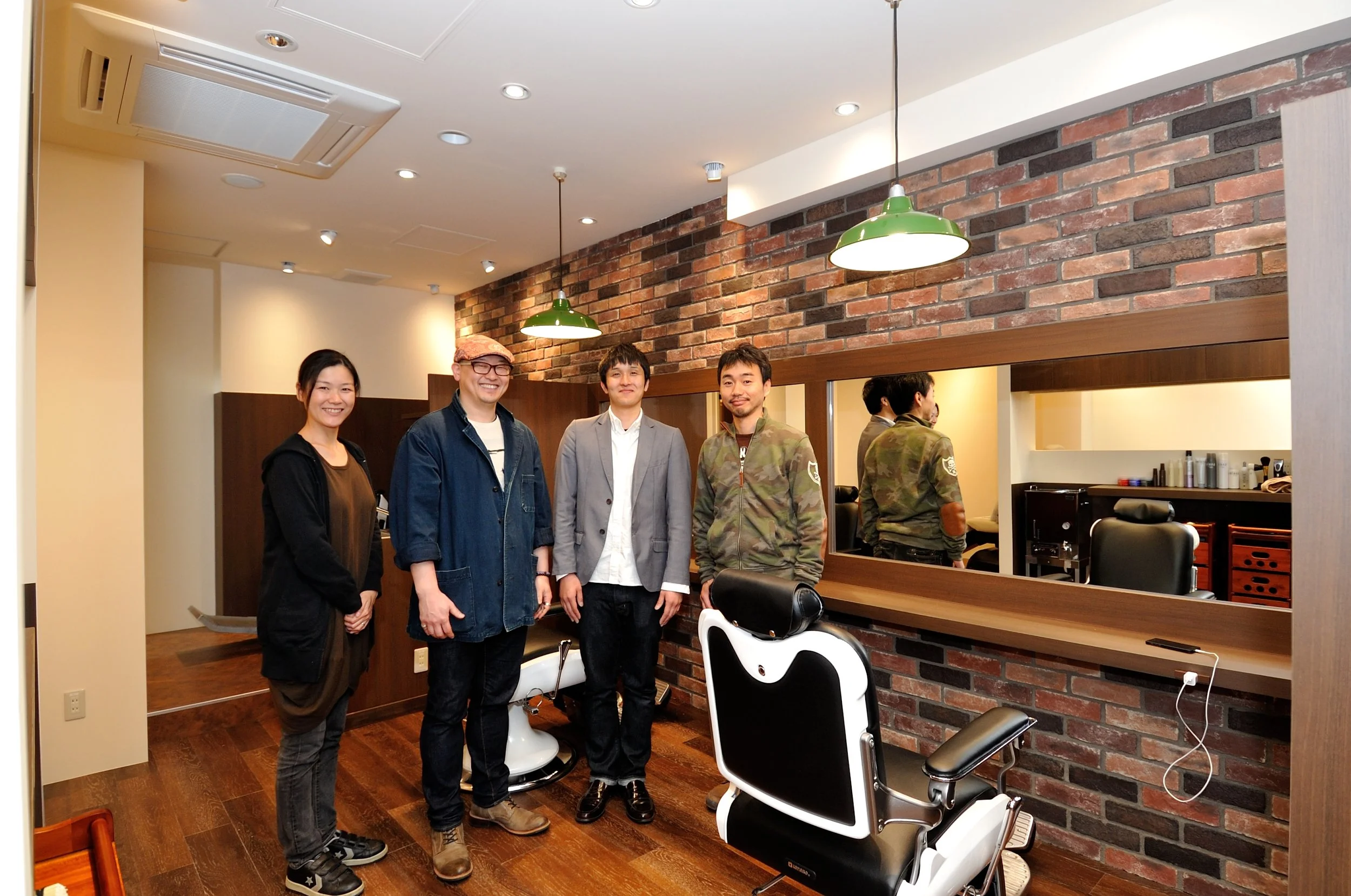 4人の男女が美容院の店内で笑顔で立っている。背景にはレンガ壁と鏡、椅子、ヘアケア用品が見える。