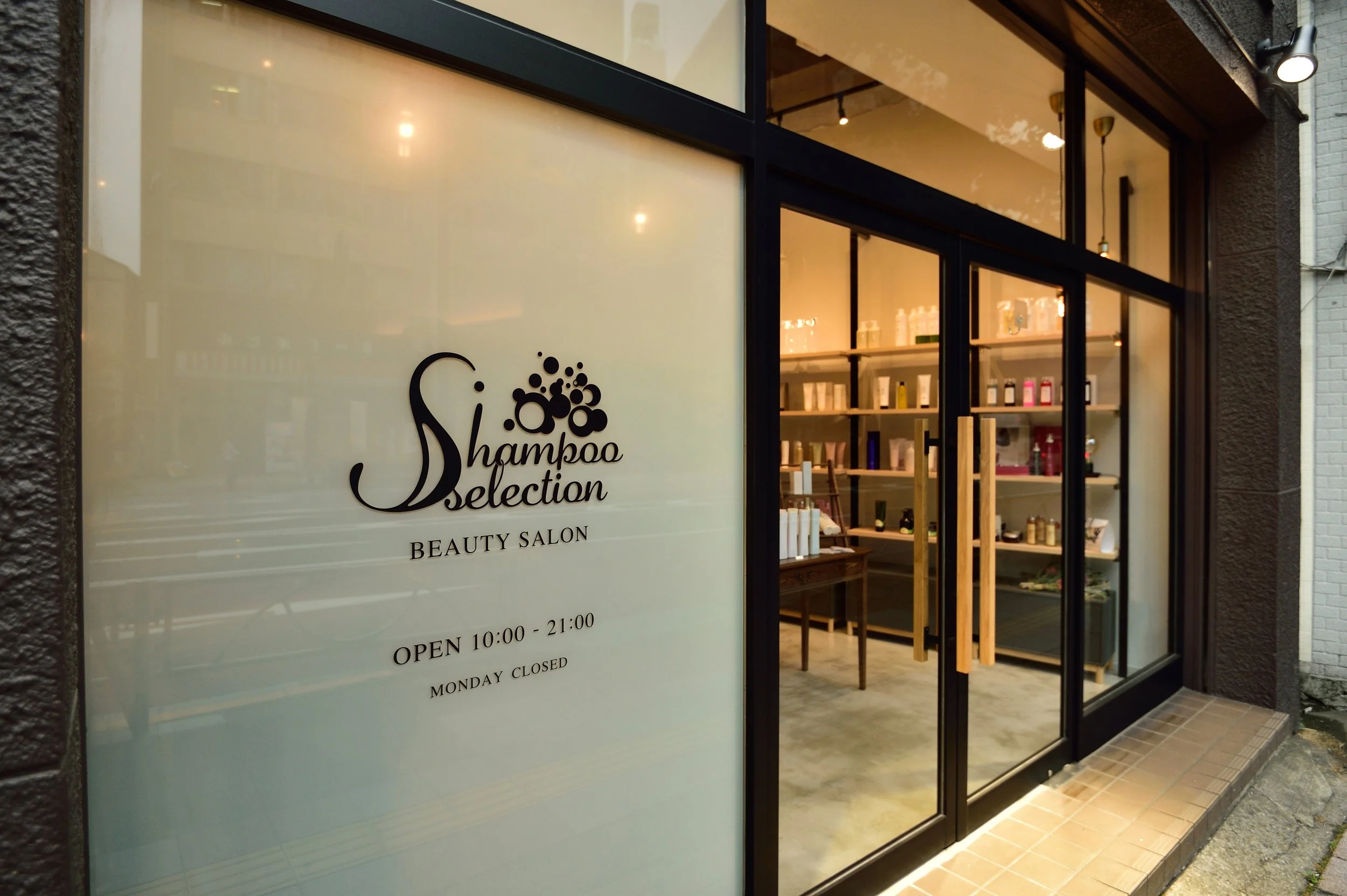 美容サロンの入り口。窓に『Shampoo Selection』と書かれ、営業時間は午前10時から午後9時まで、月曜日は休業と記載されている。店内には棚に美容用品が並んでいる。