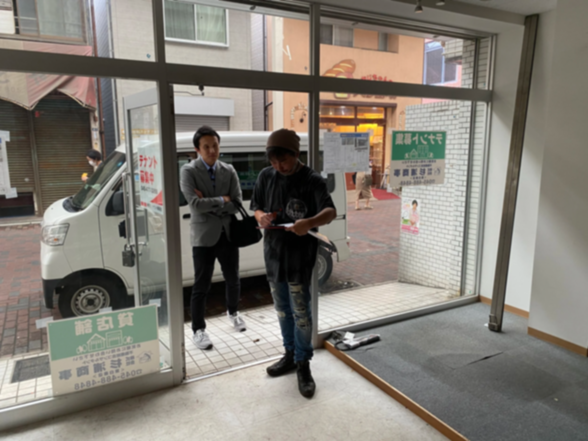 店頭で商品を確認している二人の男女と、店内から外を見る別の人がいるシーン。窓の外には通行人やバイク、自動車が見える。
