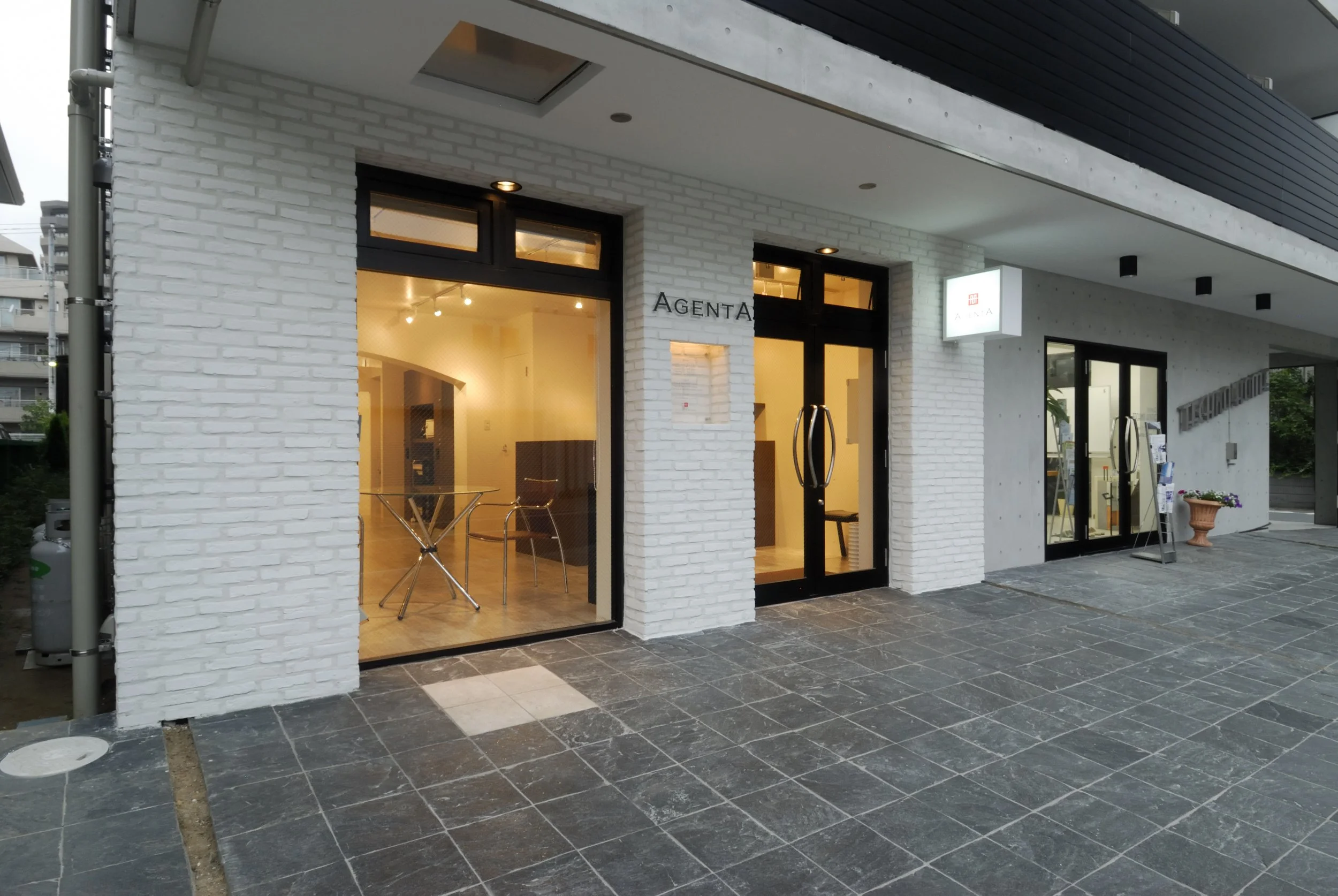 白いレンガの壁の建物のショップの入り口。ガラスのドアと窓、店内には椅子やテーブルが見える。外にはプランターとタイルの舗装がある。