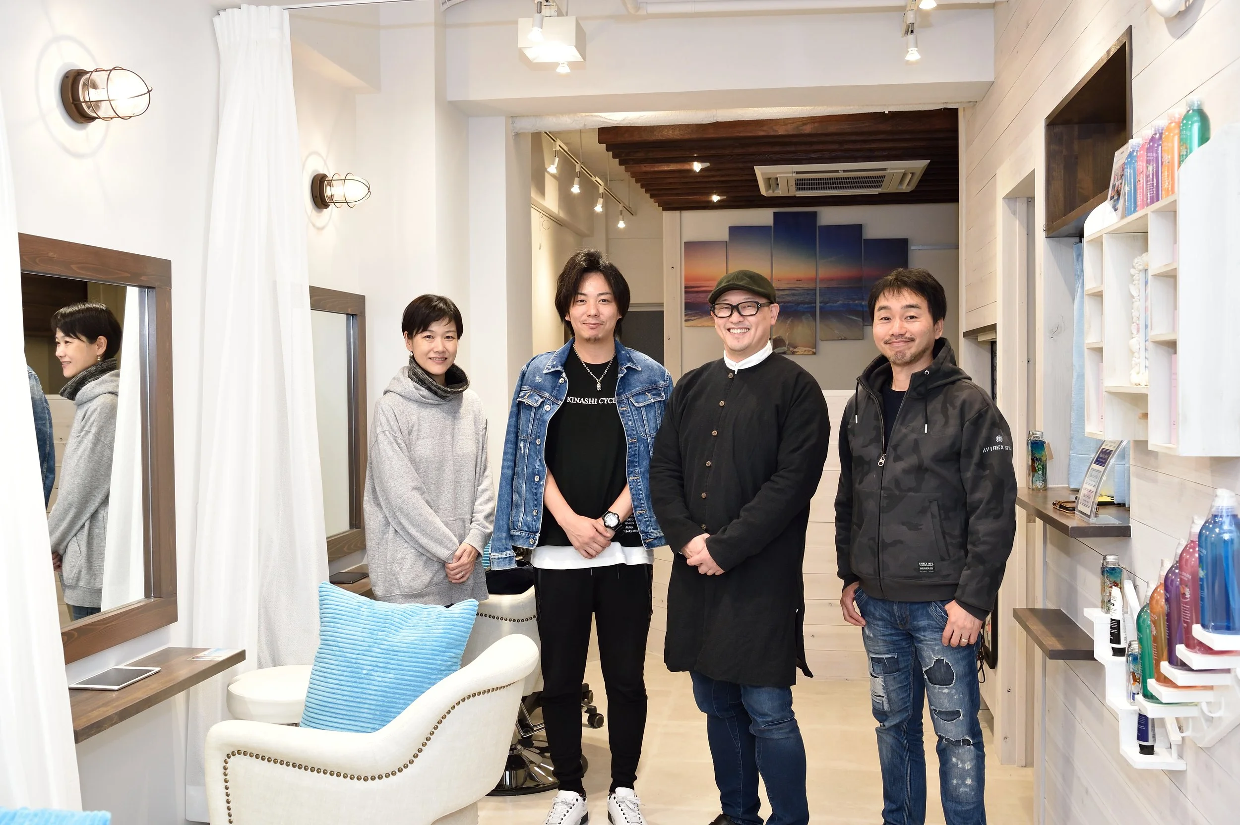 白い壁と天井の明るいインテリアに、4人の笑顔の人々が立っている。背景には海の夕焼けの絵画。左側に鏡と白いイスがあり、右側に棚とカラフルな商品が並ぶショップの一角の画像です。
