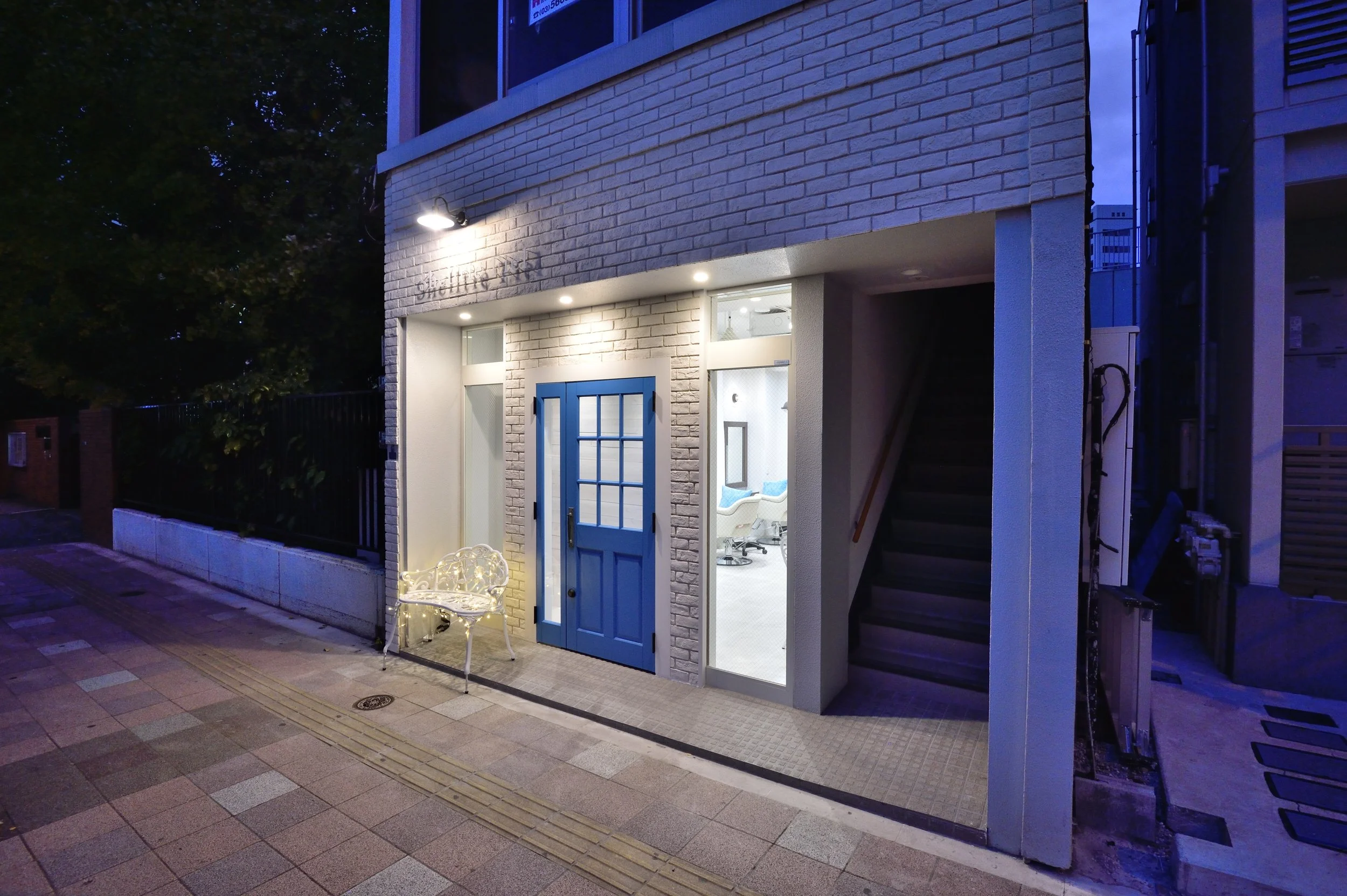 夜の建物の入り口で、青いドアと白いレンガの壁が見える。外には白い椅子とライトが置かれている。