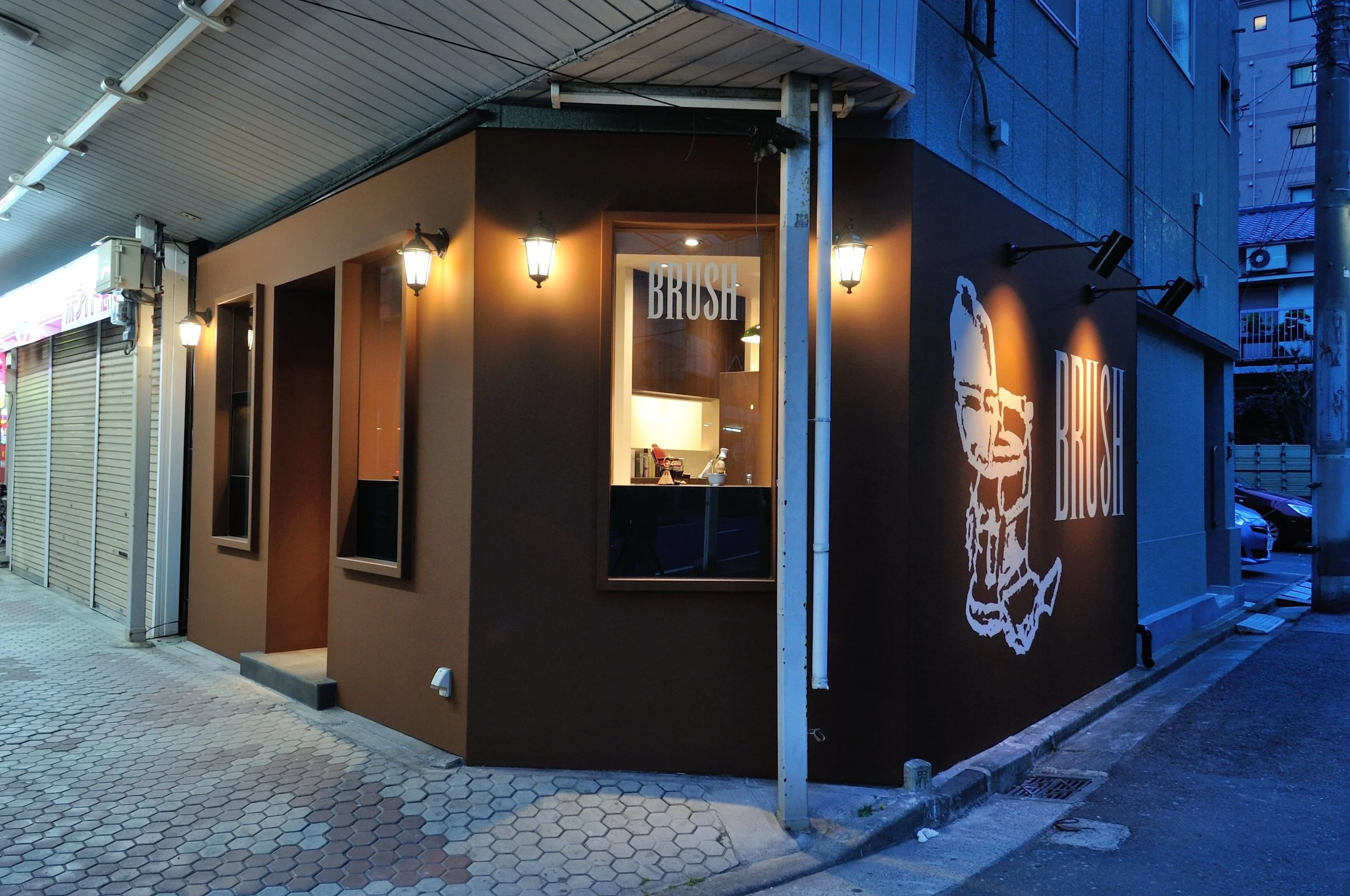 夜の街角にある理容室の外観で、暖かい照明が灯る。壁には「BRUSH」という文字と、顔のシルエットのイラストが描かれている。