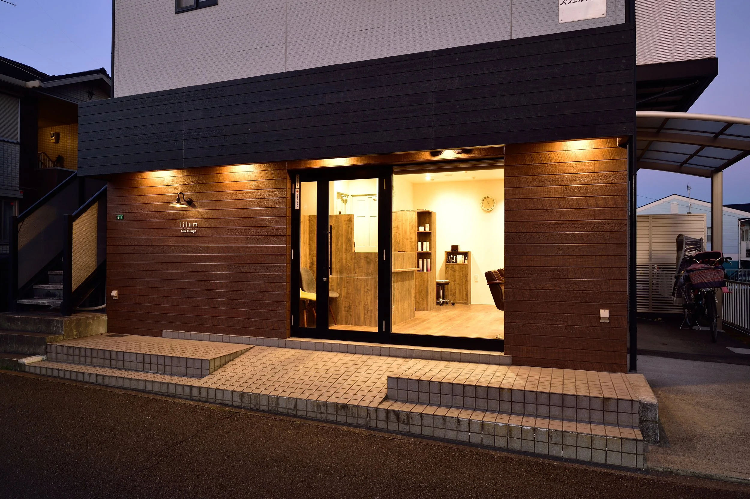 木製の外観の美容室の入口。店名はイルム ヘアラウンジと書かれている。内部は温かみのある照明と木製の家具が見える。