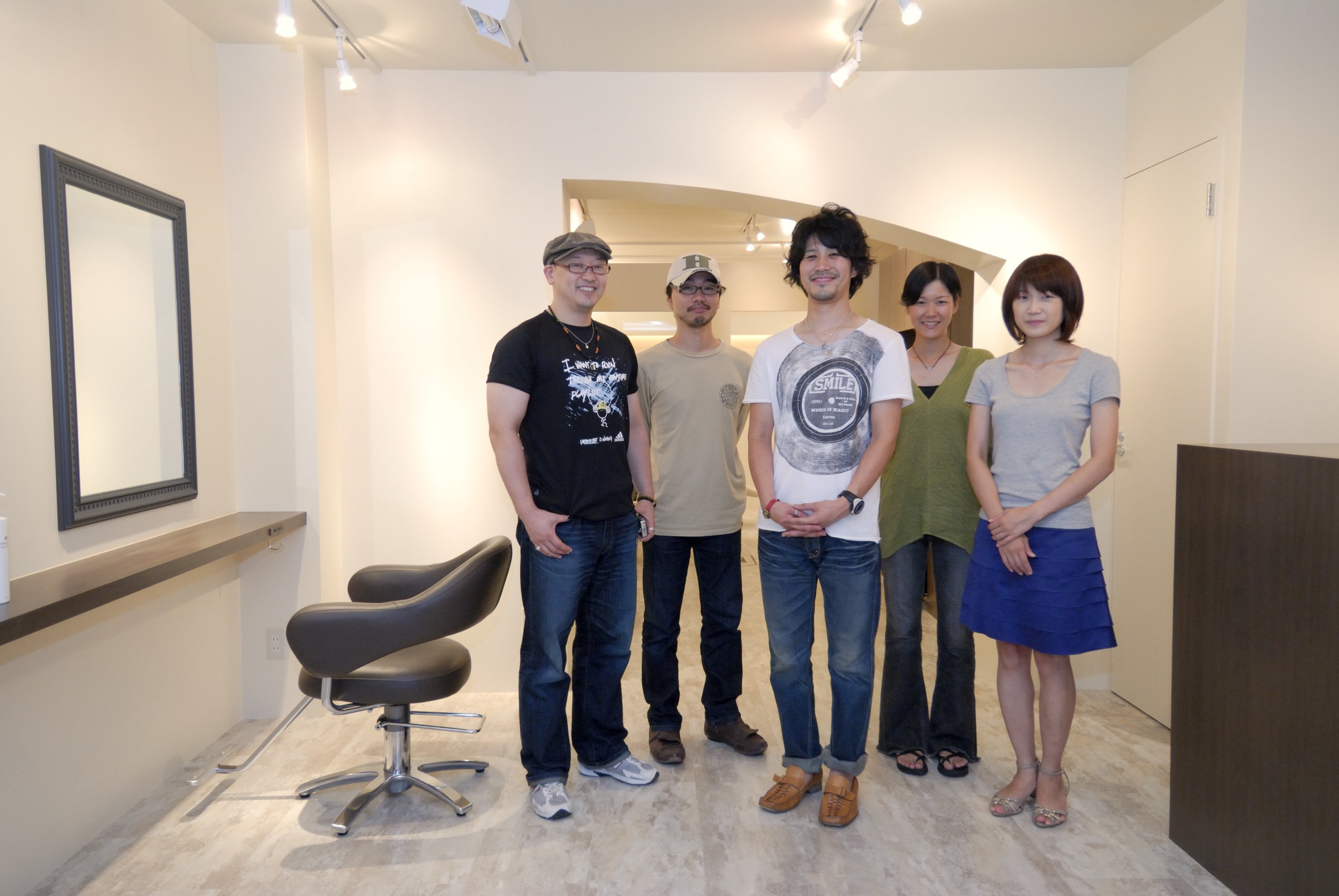 室内で5人の男女が並んで写真を撮っている。背景は白い壁といくつかのミラーや椅子が見える。