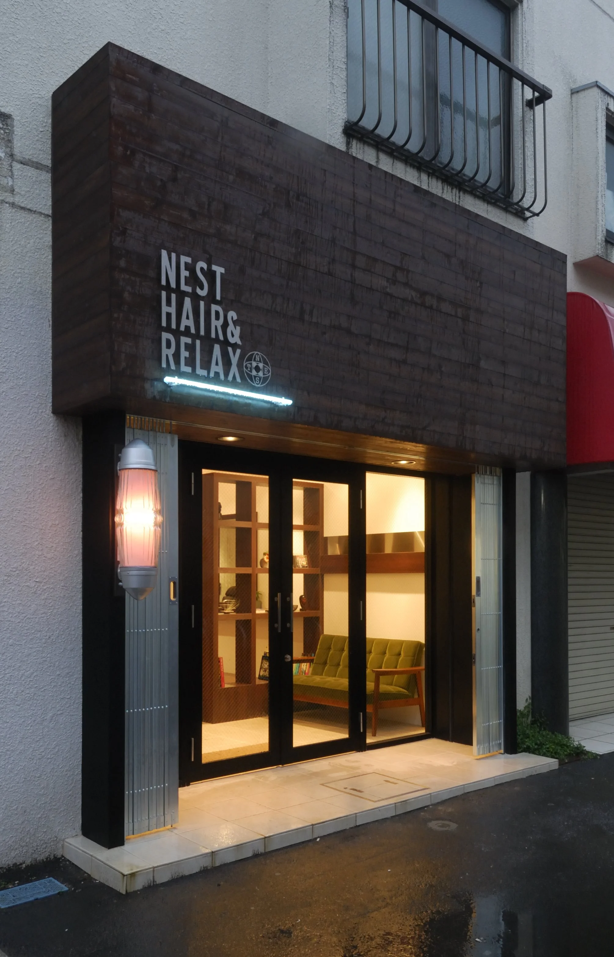店の入り口の看板に「NEST HAIR & RELAX」と書かれている。ガラスの引き戸の内側に緑色のソファと本棚が見える。