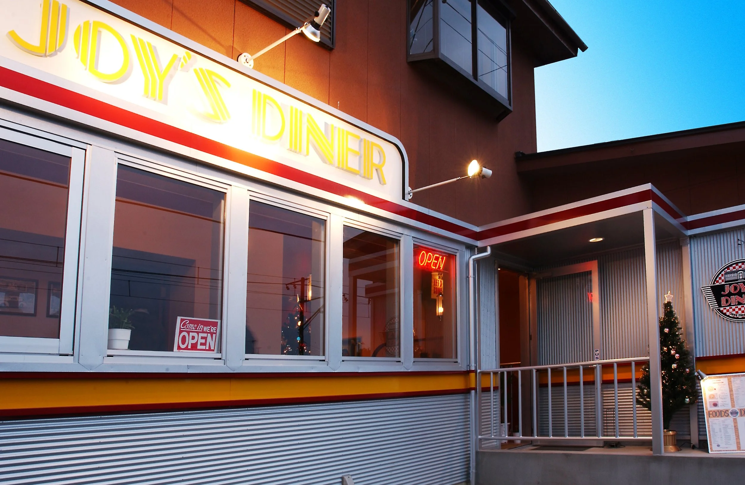 夕暮れ時のレストランの外観で、入り口に『JODY'S DINER』と黄色のネオンサインと赤色の『OPEN』の表示がある。大きな窓やドア、メニューが置かれた立て看板も見える。