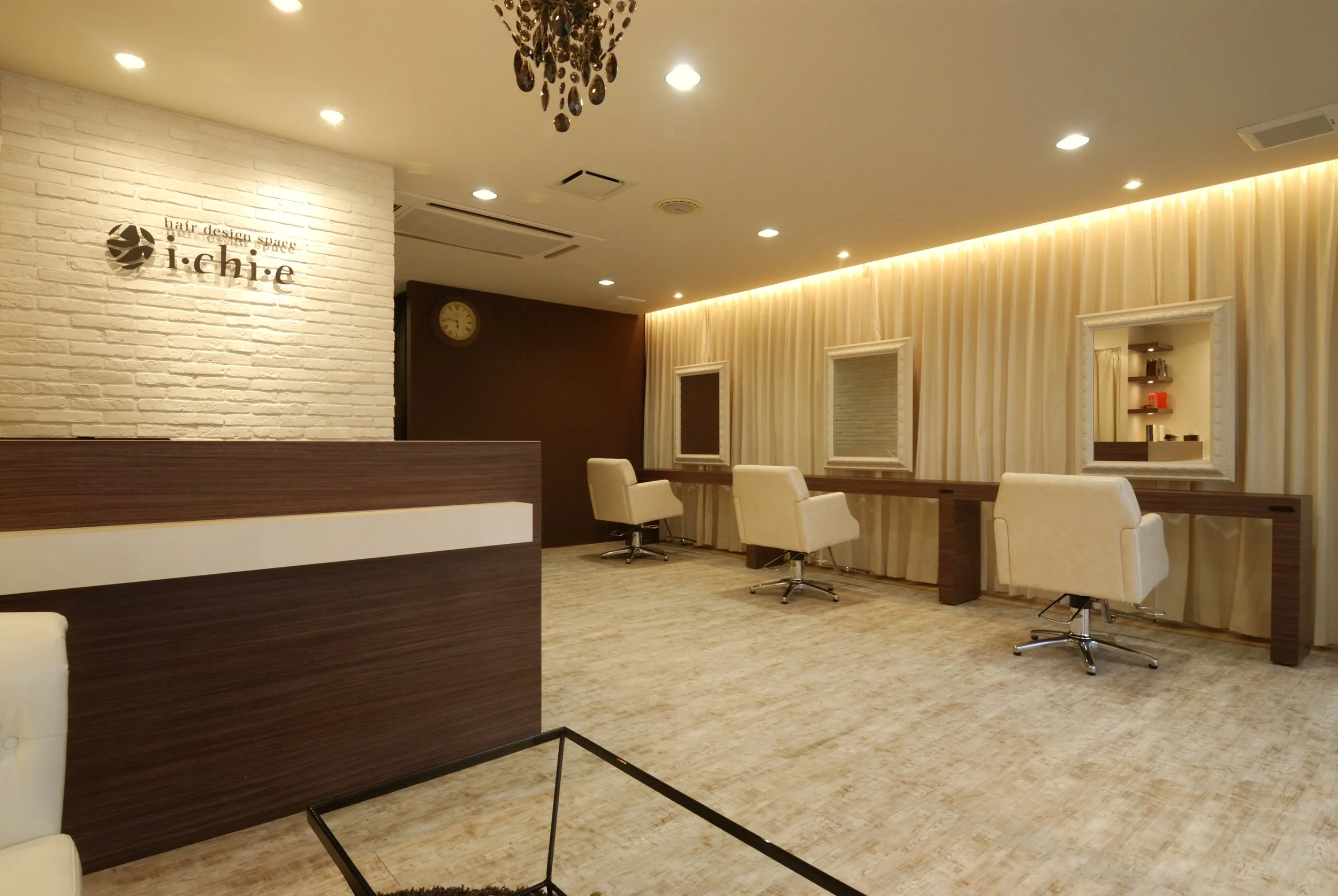 白いレンガの壁に「i-chi:e hair design space」のサインとロゴ。木製の仕切りと白い椅子が並ぶ美容院の待合スペース。鏡と飾り棚が壁に設置され、暖色の照明が温かい雰囲気を作り出している。