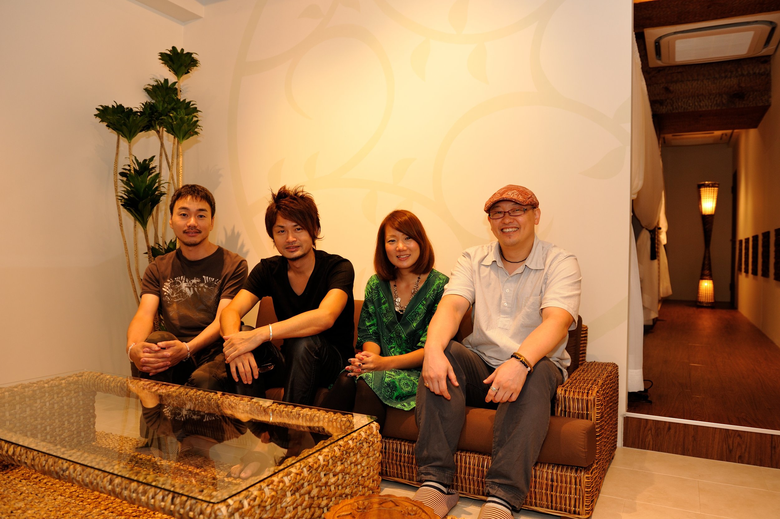 木製のソファに座っている4人の人々の写真。背景には観葉植物と暖色系の照明が見える。