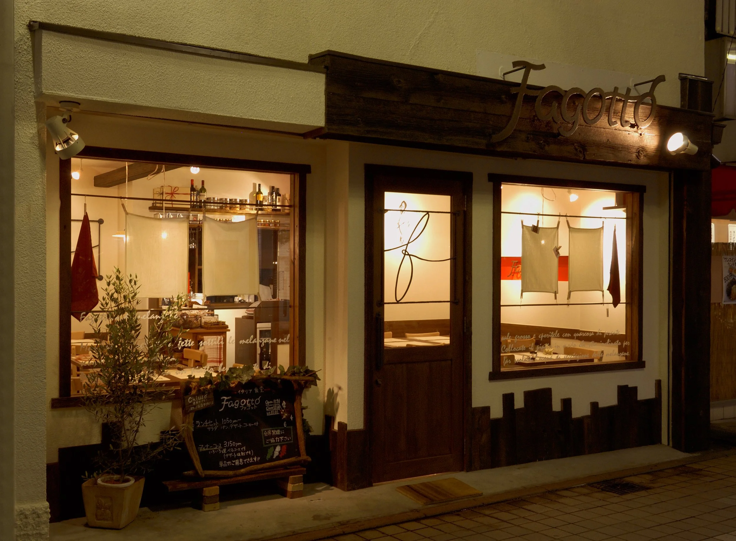 木製の看板に書かれた「Toscotto」の文字があるイタリアンレストランの店舗外観。窓の内側に照明と壁飾りが見え、黒板メニューも設置されている。」}】}
