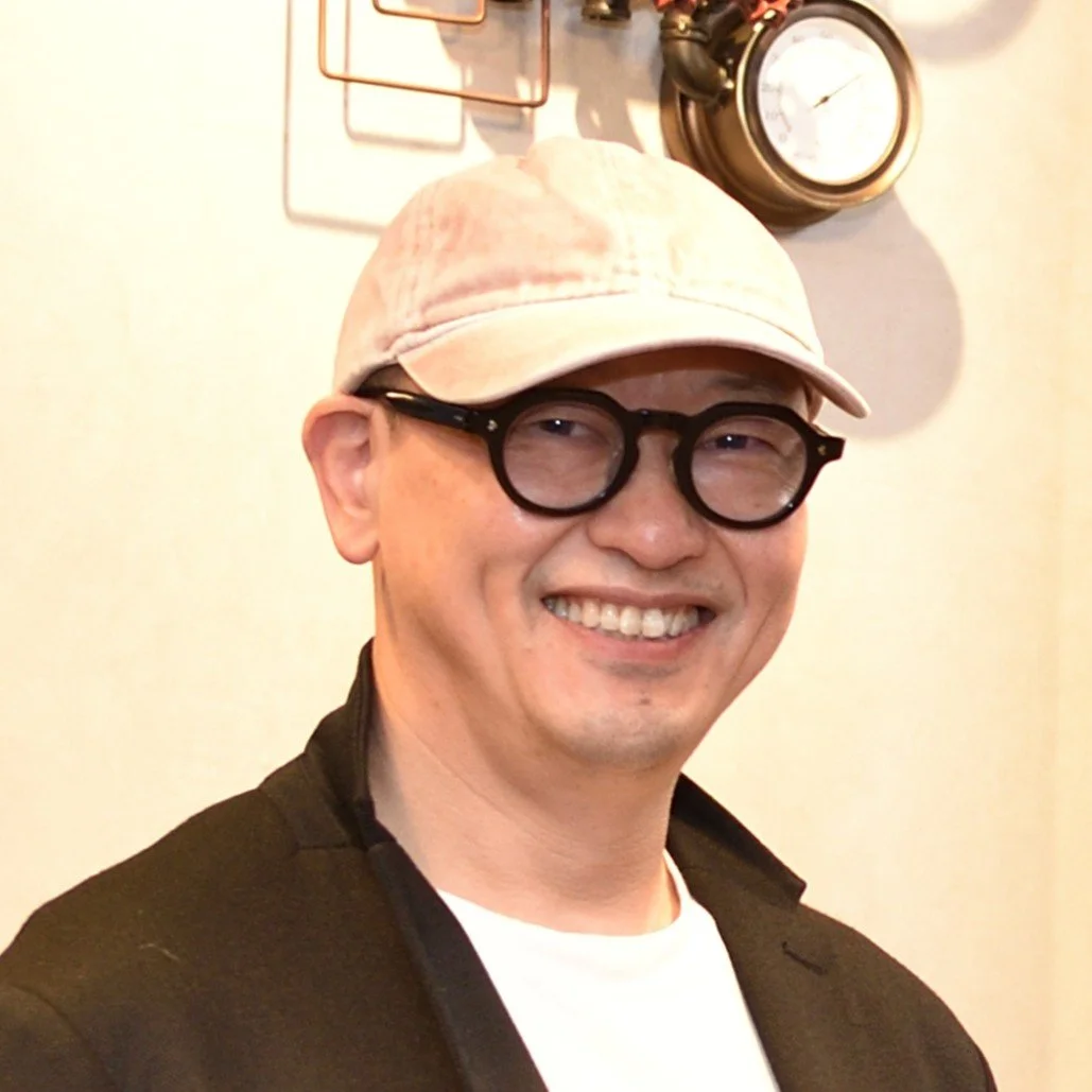 笑顔の男性、白いキャップと丸い眼鏡を着用、背景に時計と装飾物がある。