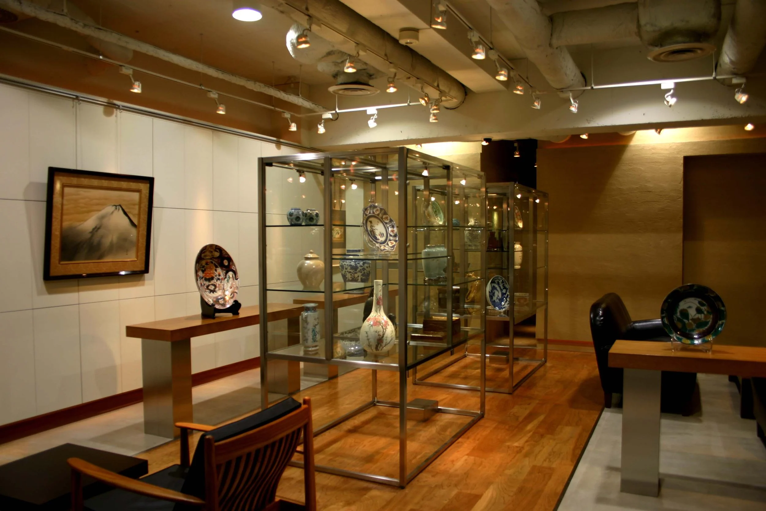 展示棚に陶器の花瓶や皿が飾られている室内の風景。壁には絵画が掛かっている。木製の椅子が前景にある。照明は天井から多くのスポットライトで照らされている。