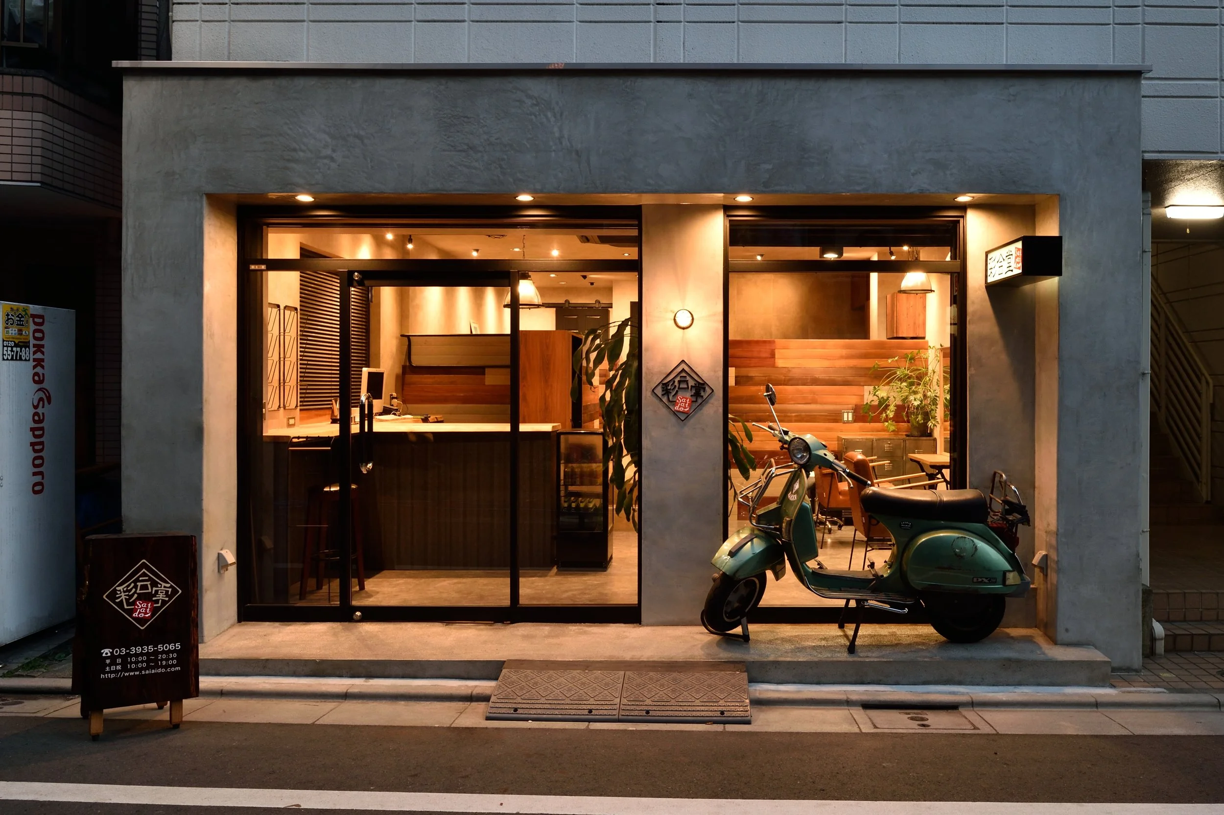 夜の東京の小さなレストランの正面。大きなガラス窓越しに木製の内装と暖色の照明が見える。入口の左側に黒い看板とバイクと自転車が駐輪されている。