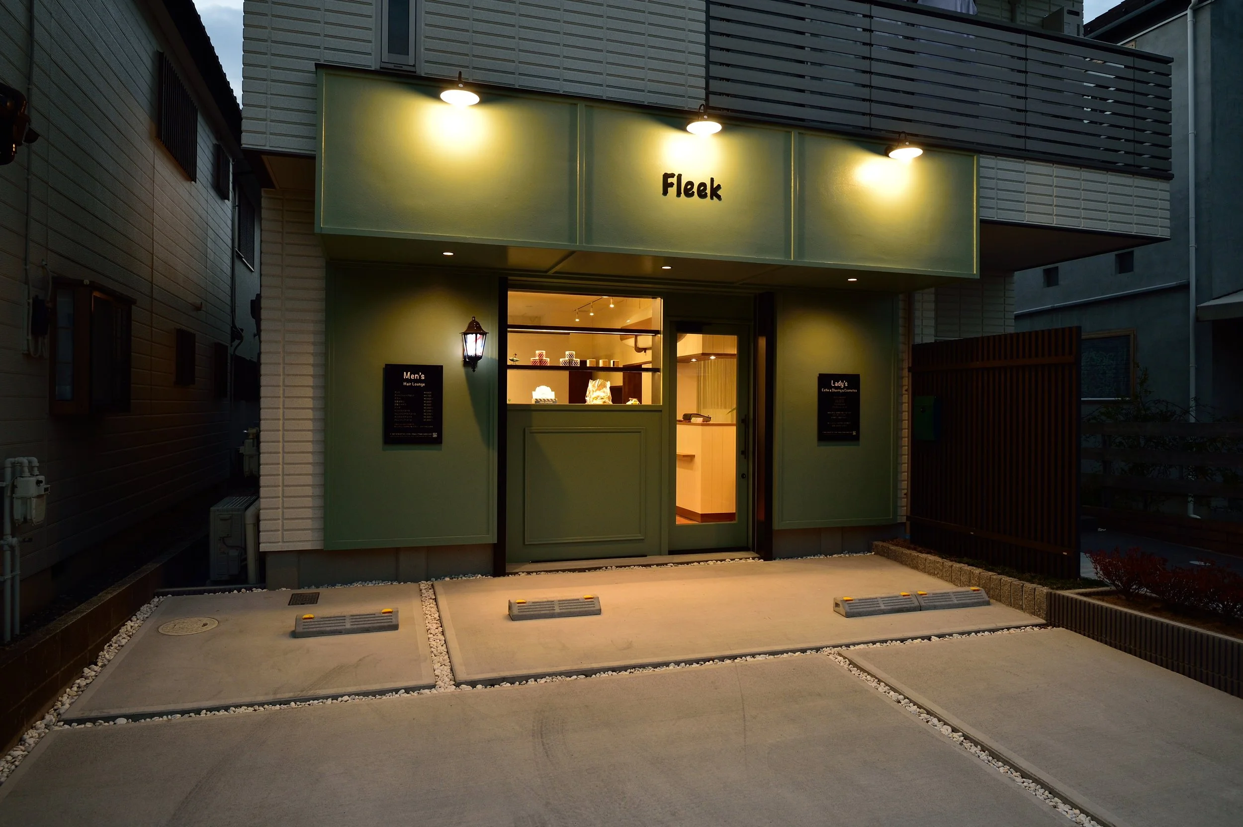 緑色の外観の小さな店の正面、名前はFleekと書かれている。照明が灯り、店内が見える。駐車スペースと小さな石の装飾が前に配置されている。周囲は都市の建物に囲まれている。