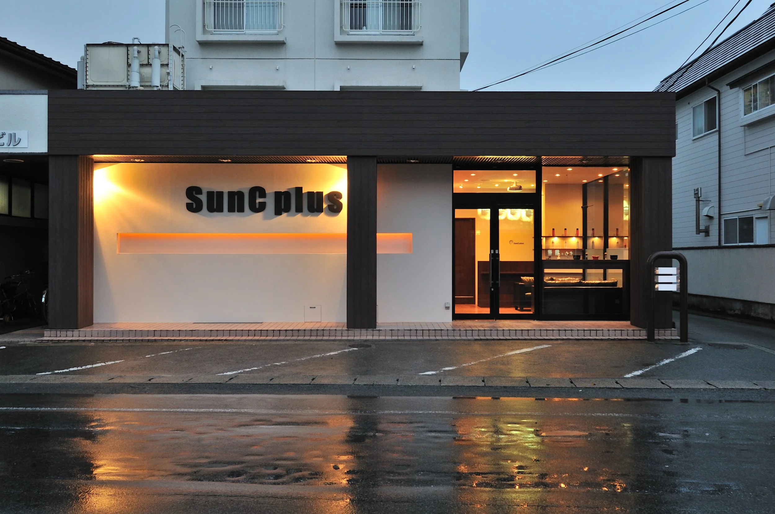 夜の街並み、店舗の入り口に暖かい光とサイン『SunC plus』の文字が見える。雨で濡れた道路と反射する光が印象的な都市の景色。
