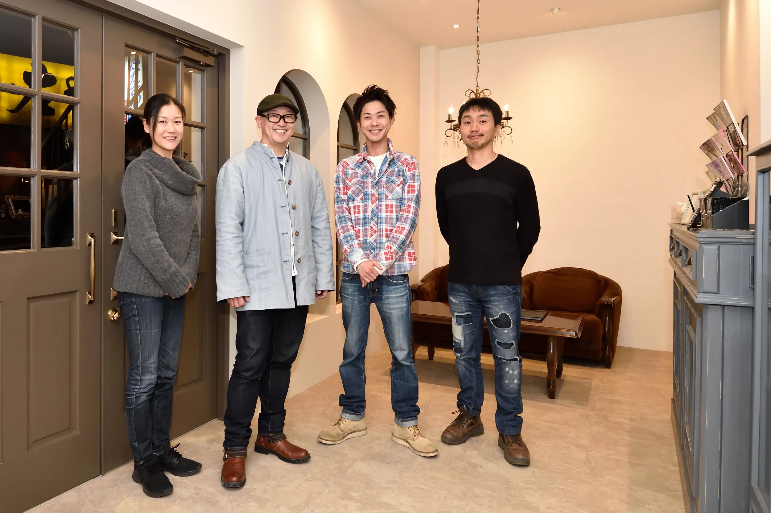 カメラに向かって笑顔で立っている5人の若い大人たち。室内の暖かい照明と暖炉が見えるリラックスした雰囲気の部屋。左側に扉と窓、右側に本や書類が並ぶ棚がある。