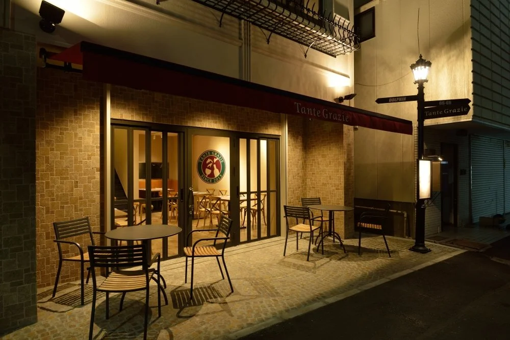 夜の街のレストランの外観。テラス席に椅子とテーブルがあり、店の入り口には看板と暖炉灯がある。