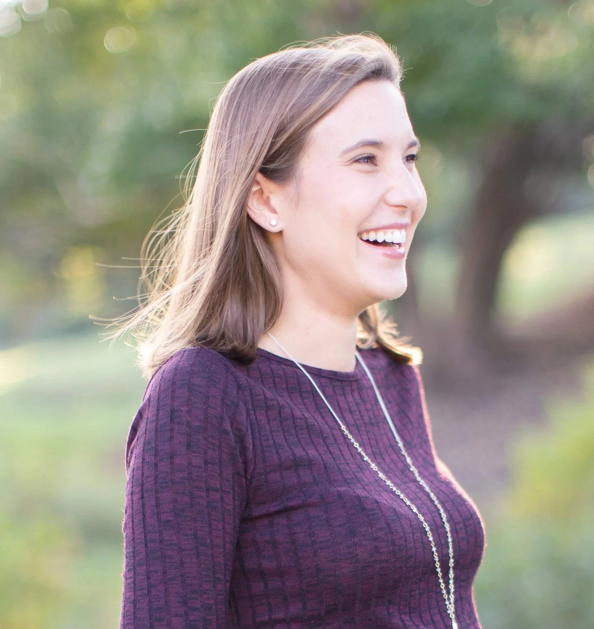 A young woman with brown hair smiling outdoors, wearing a purple long-sleeve shirt and jewelry.
