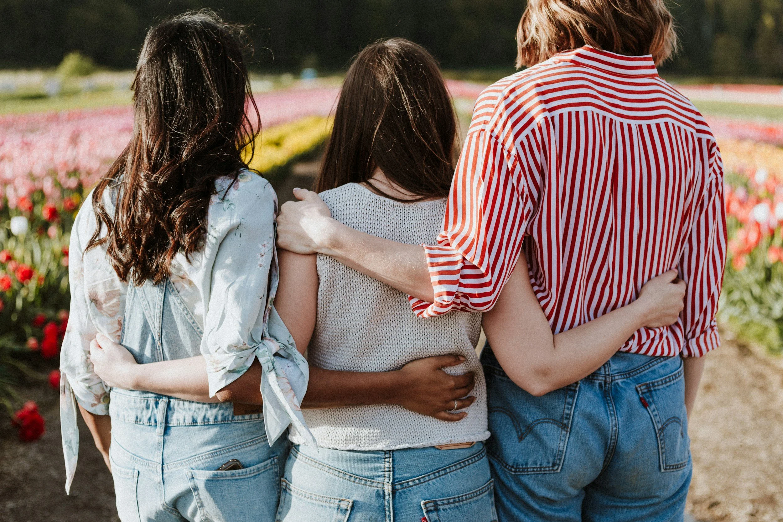 Three women standing together in a flower field, with their arms around each other, facing away from the camera.