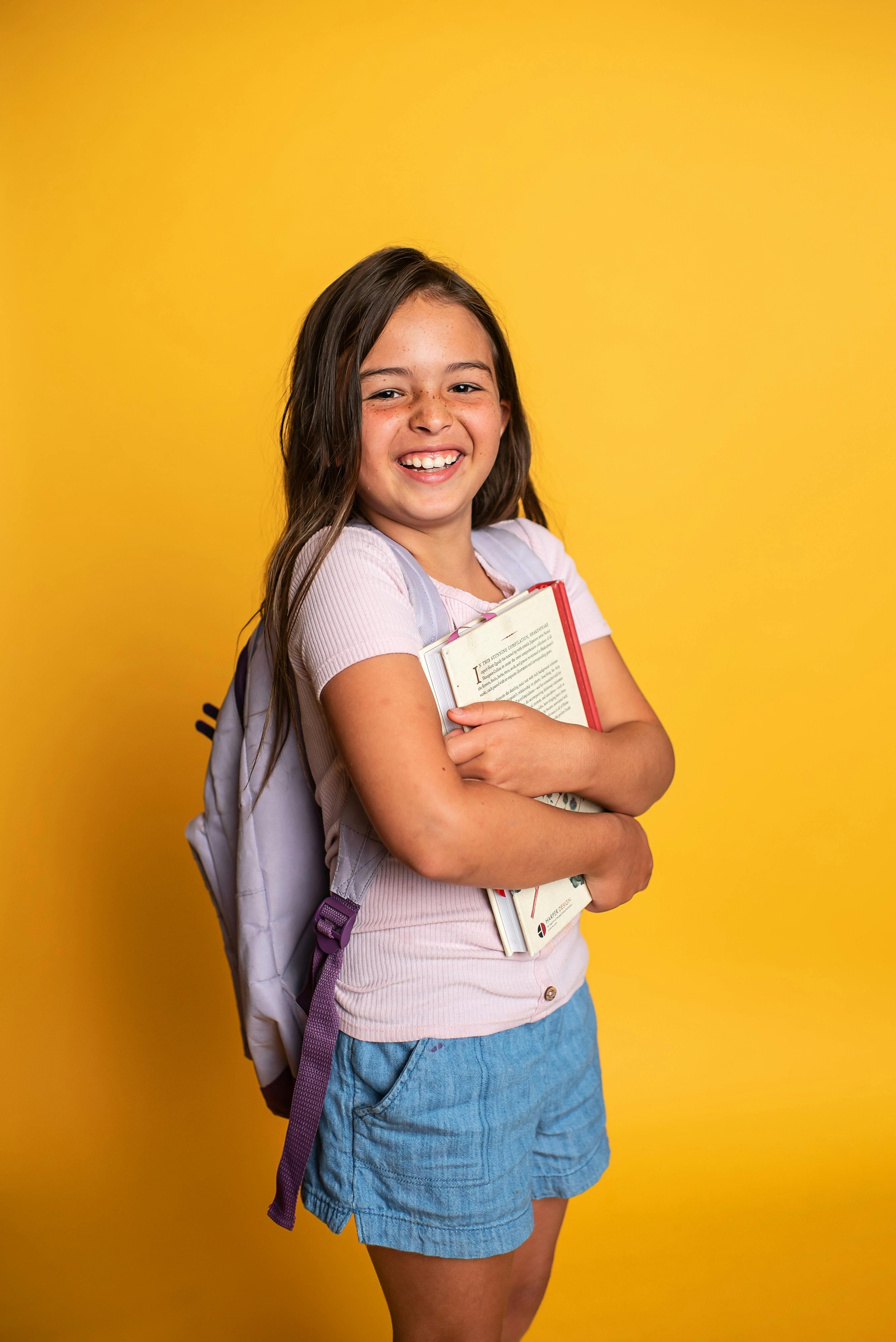Smiling school-age girl with a backpack hugging her books against a bright yellow background