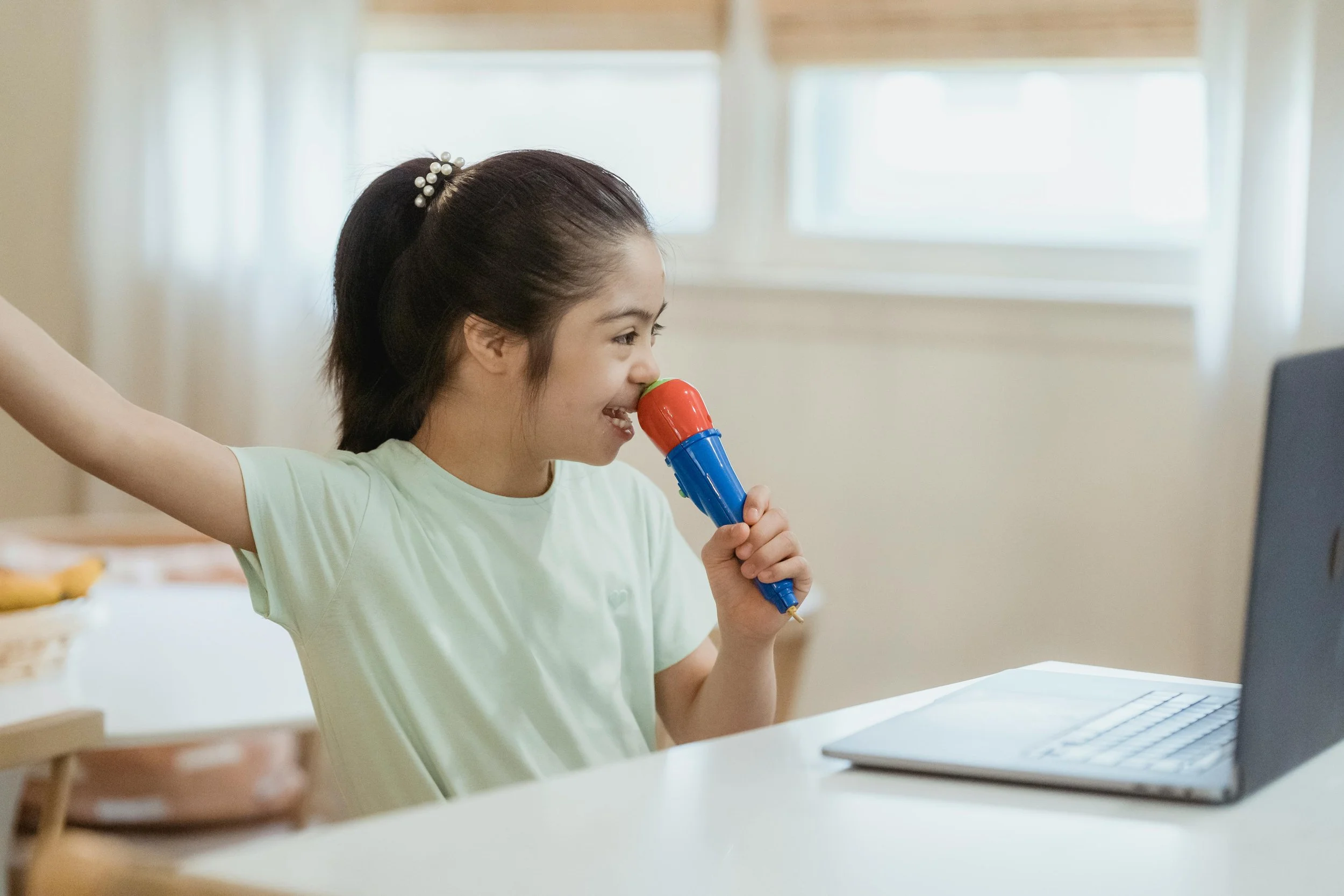 Young girl with dark hair in a ponytail holding a toy microphone and looking at a laptop.