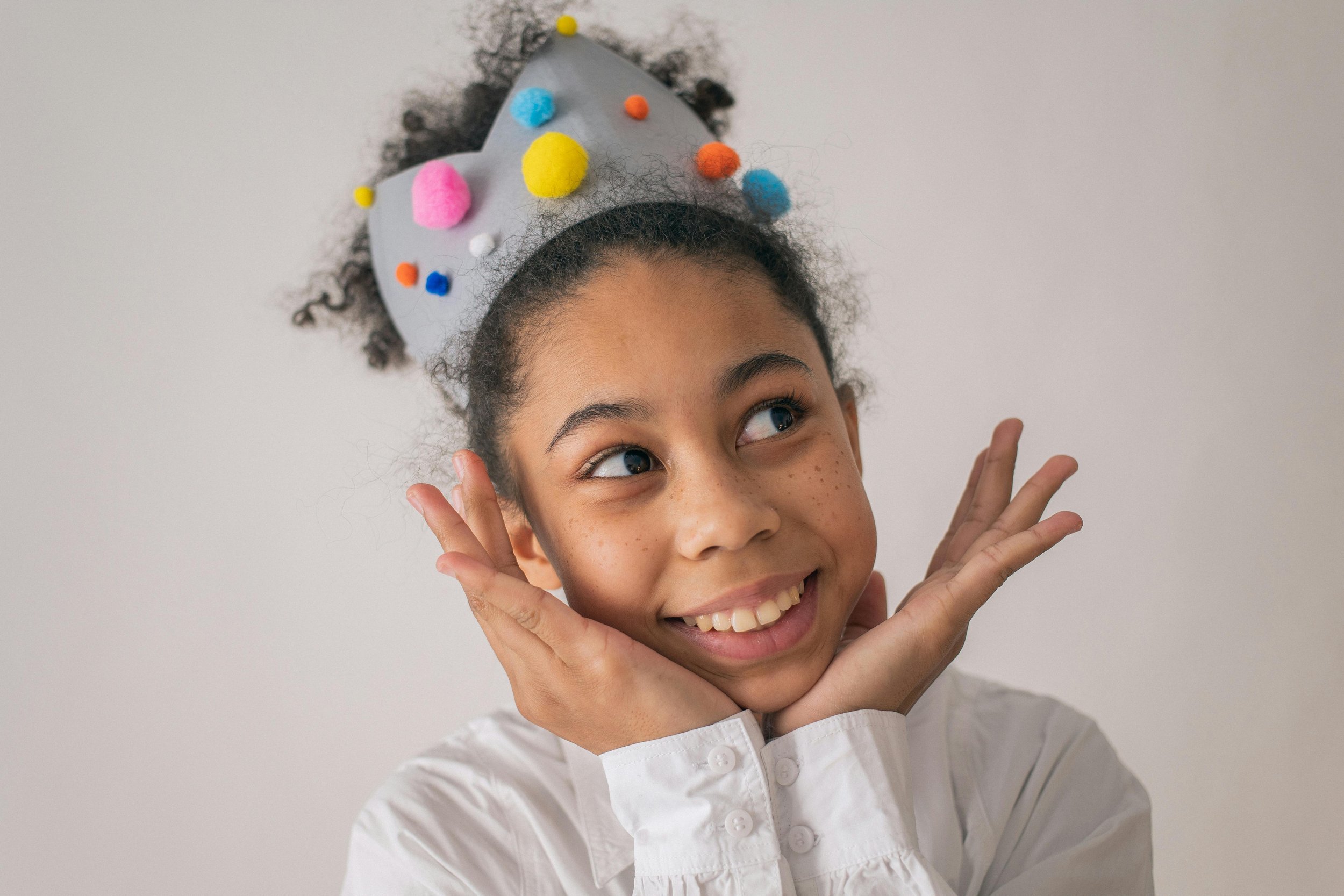 A young girl with curly hair, freckles, and bright eyes smiling and posing with her hands near her face, wearing a colorful felt hat decorated with pom-poms.
