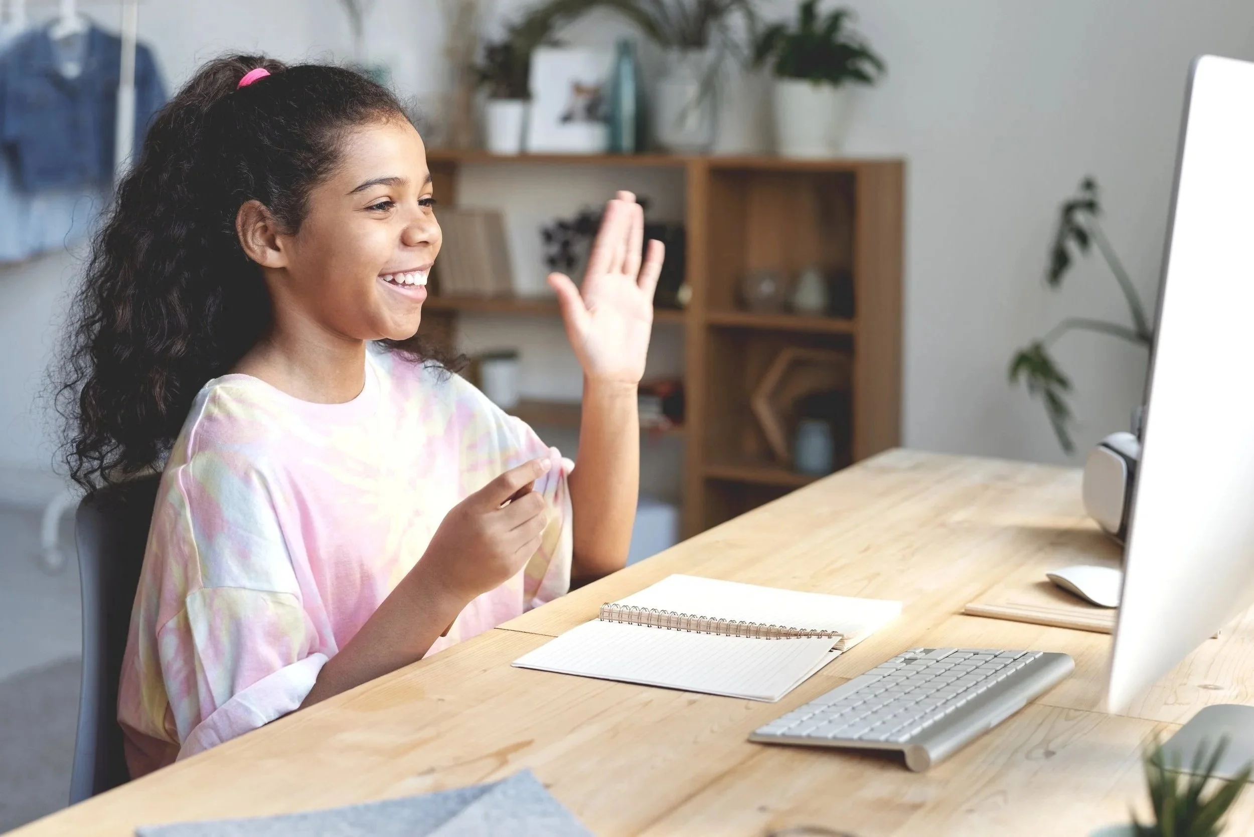 A young girl with curly hair, smiling and waving while participating in a video call on a desktop computer in a well-lit room.