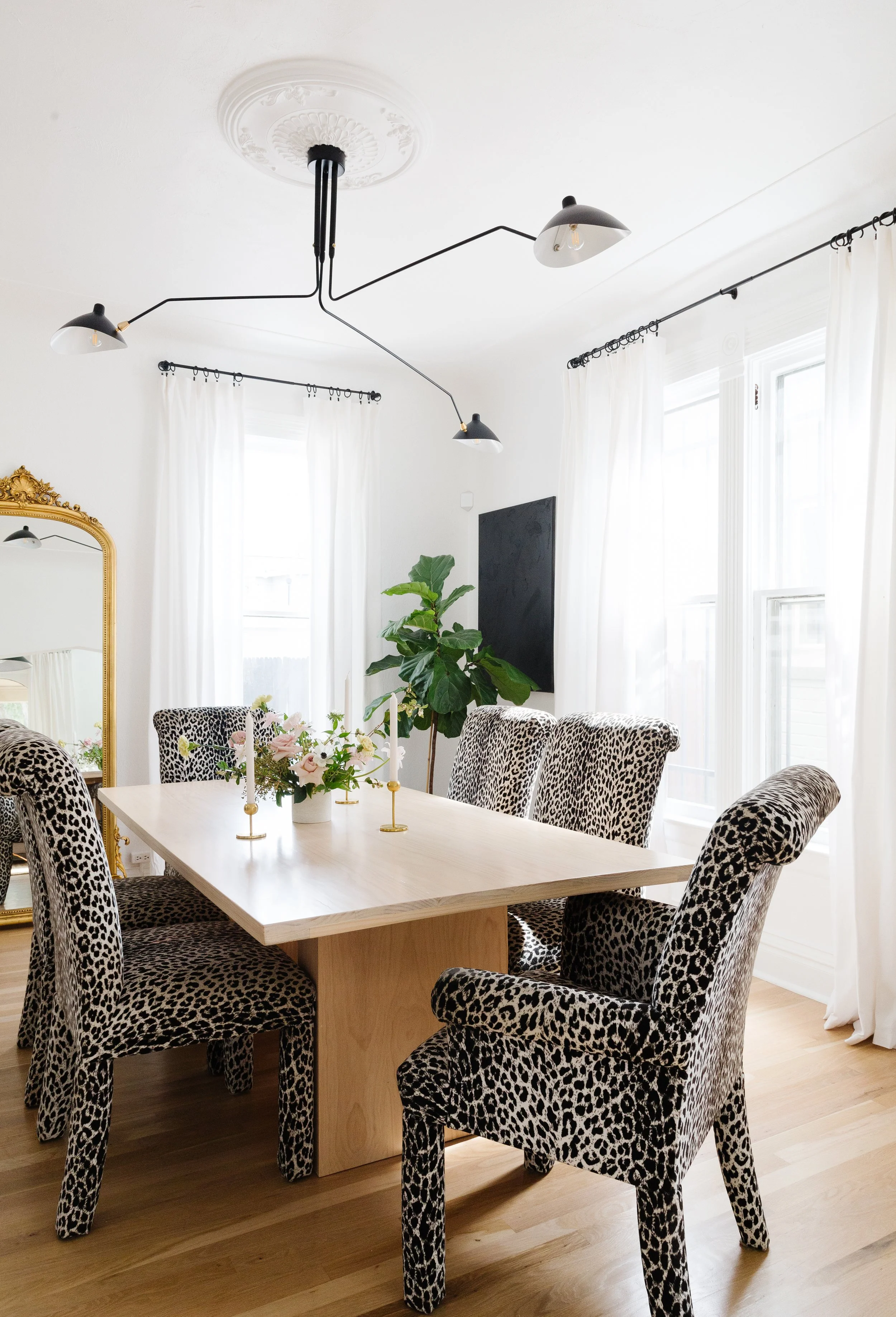 A dining room with a light wood table surrounded by six leopard print upholstered chairs, a large mirror with a gold frame on the left, white curtains on the windows, a potted plant in the corner, a black chalkboard on the wall, and a modern black ceiling light fixture.