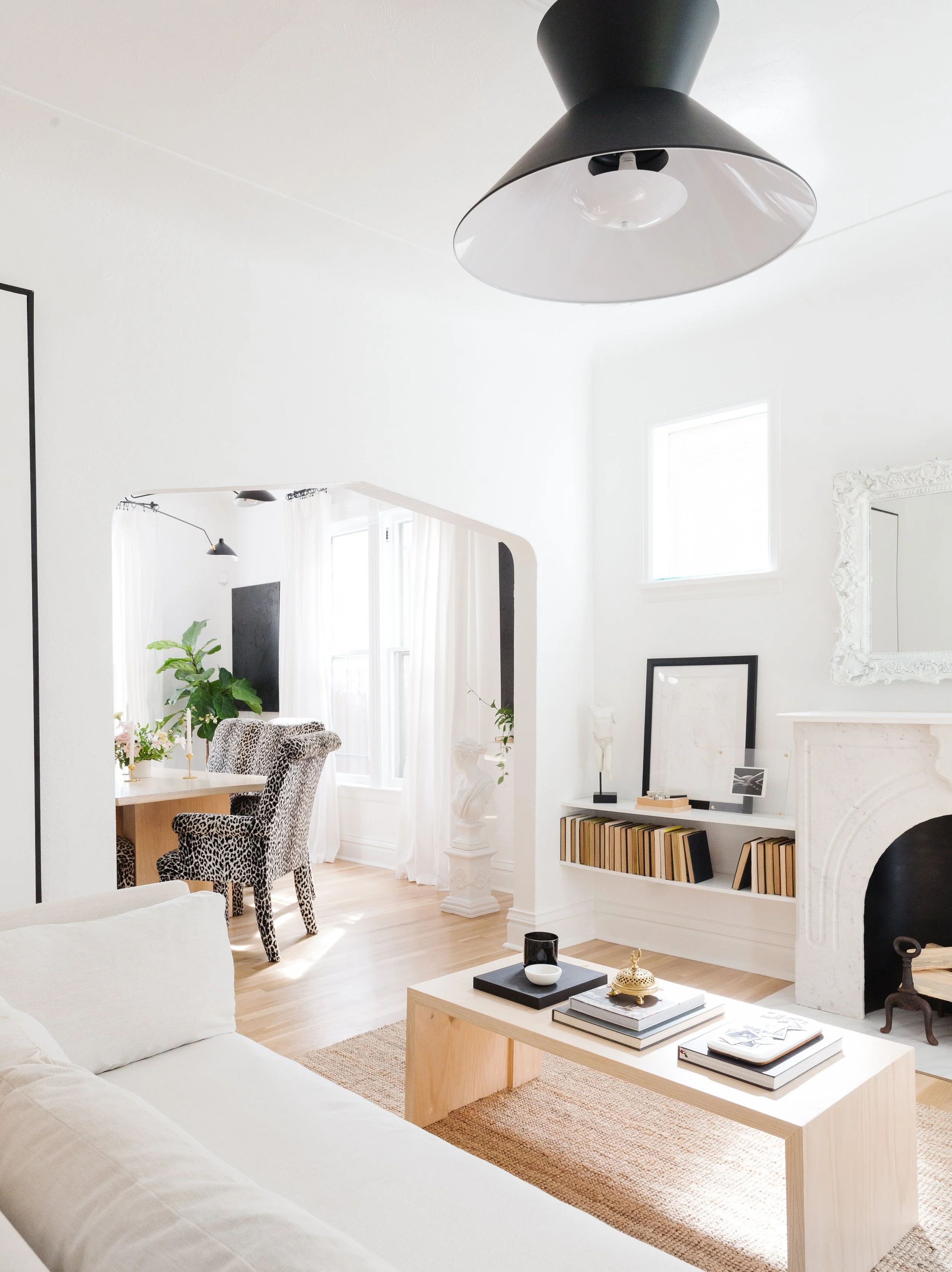 Bright living room with white walls, wooden flooring, modern decor including a white sofa, a wooden coffee table with books and decorative items, a fireplace with a mirror above, and a small bookcase with books. In the background, a dining area with a table and leopard print chairs, large windows, and white curtains.