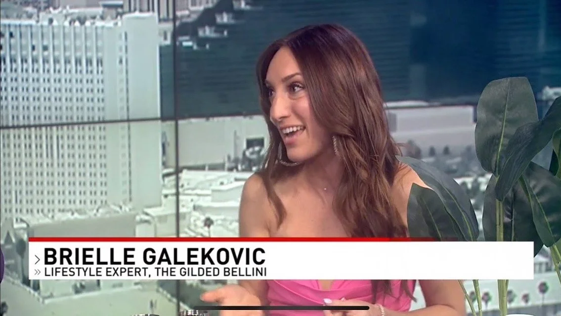 A woman with long brown hair wearing a pink top, smiling and talking during a TV interview. Behind her are city buildings and large green houseplants.