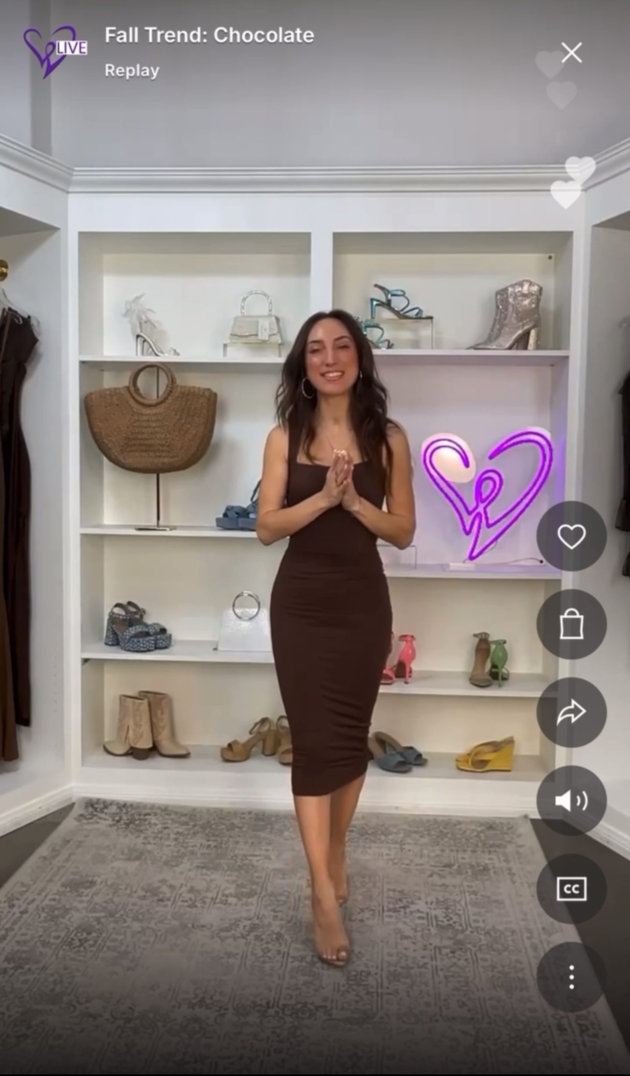 A woman standing in front of a white display shelf filled with handbags and shoes, wearing a brown dress and heels. There is a neon sign of a heart with a figure inside on the right side of the shelf.