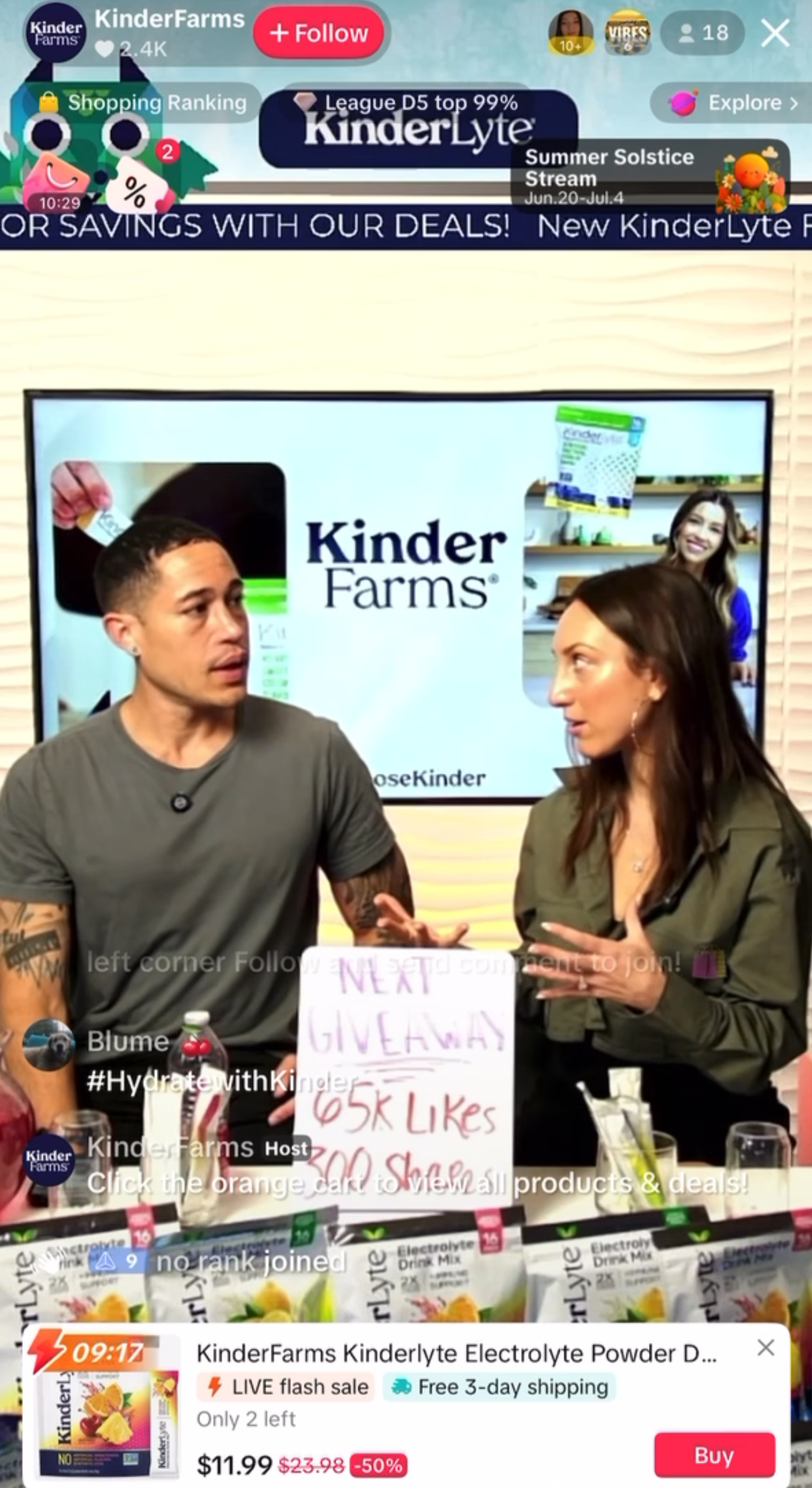 Two women are at a KinderFarms booth, discussing electrolyte powder. Behind them is a large screen displaying the KinderFarms logo. One woman is holding a sign that says 'Next Giveaway 65K Likes 30 Shop'. On the table are KinderLyte electrolyte drink