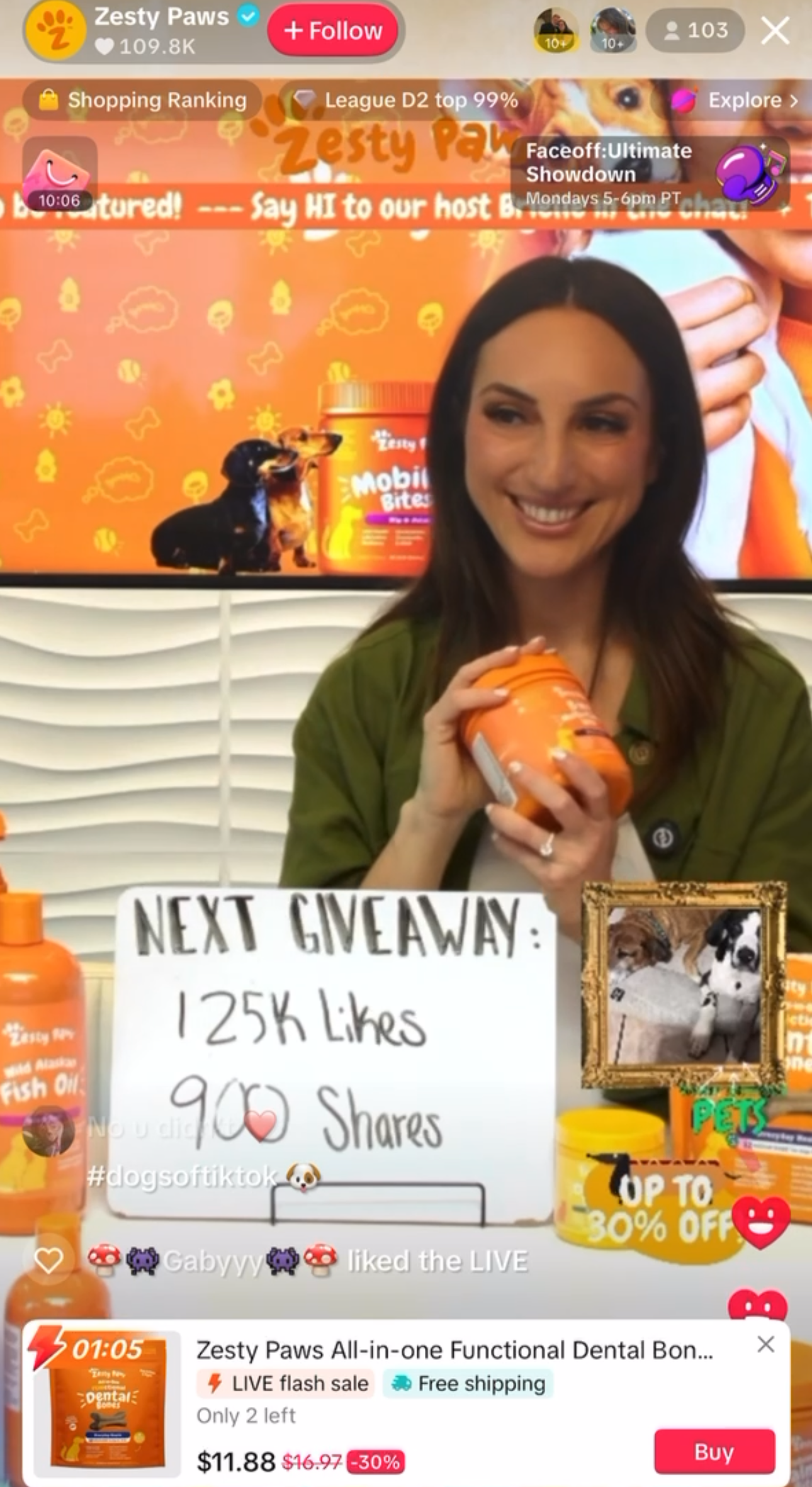 A woman holding a container of Zesty Paws pet supplement, smiling in front of a bright orange promotional display. The display features cartoon images of dogs, a jar of the supplement, and a framed photo of two puppies. There's a sign advertising a g