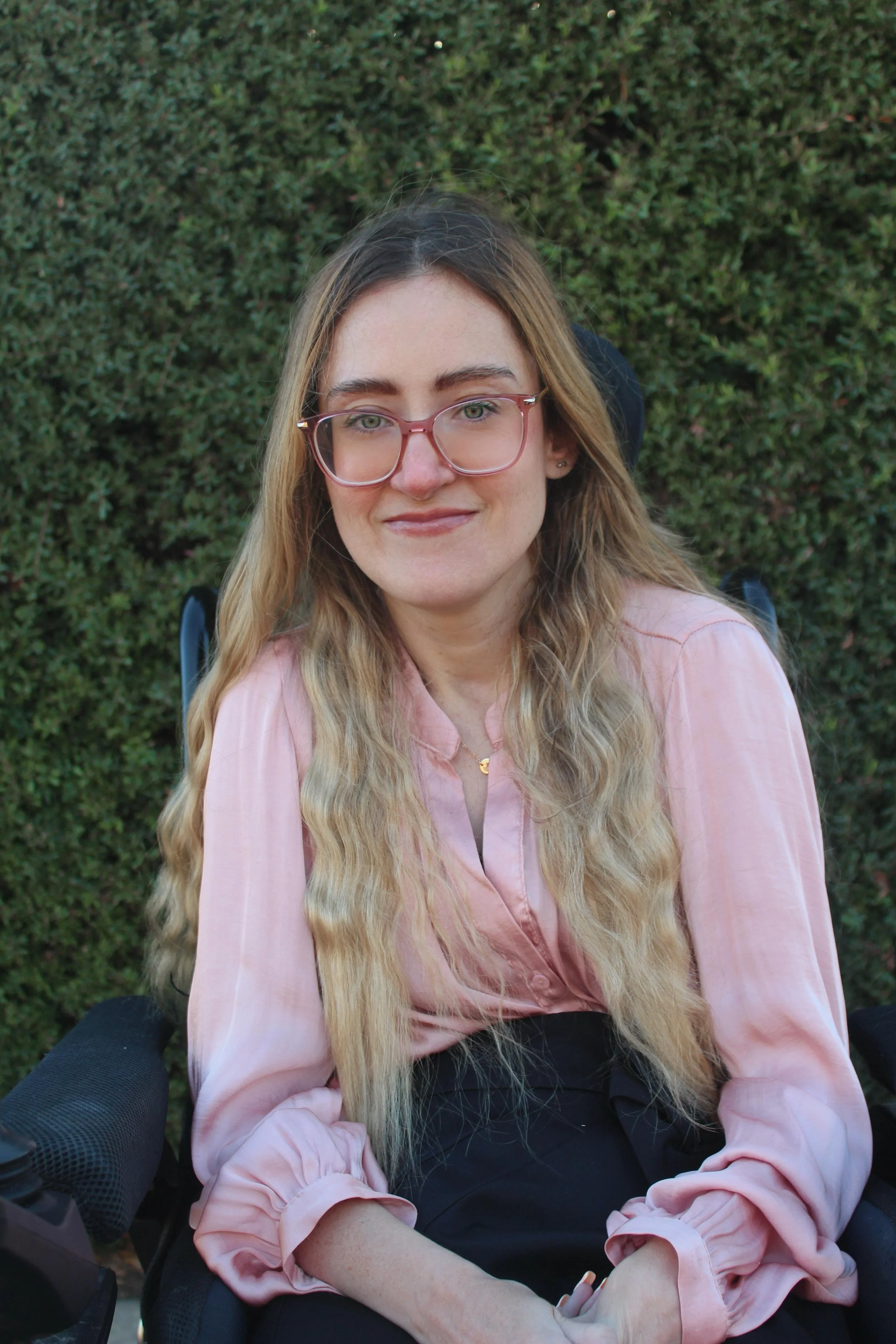 A woman with long wavy blonde hair, wearing glasses and a pink blouse, sitting in a wheelchair outdoors with green bushes in the background.